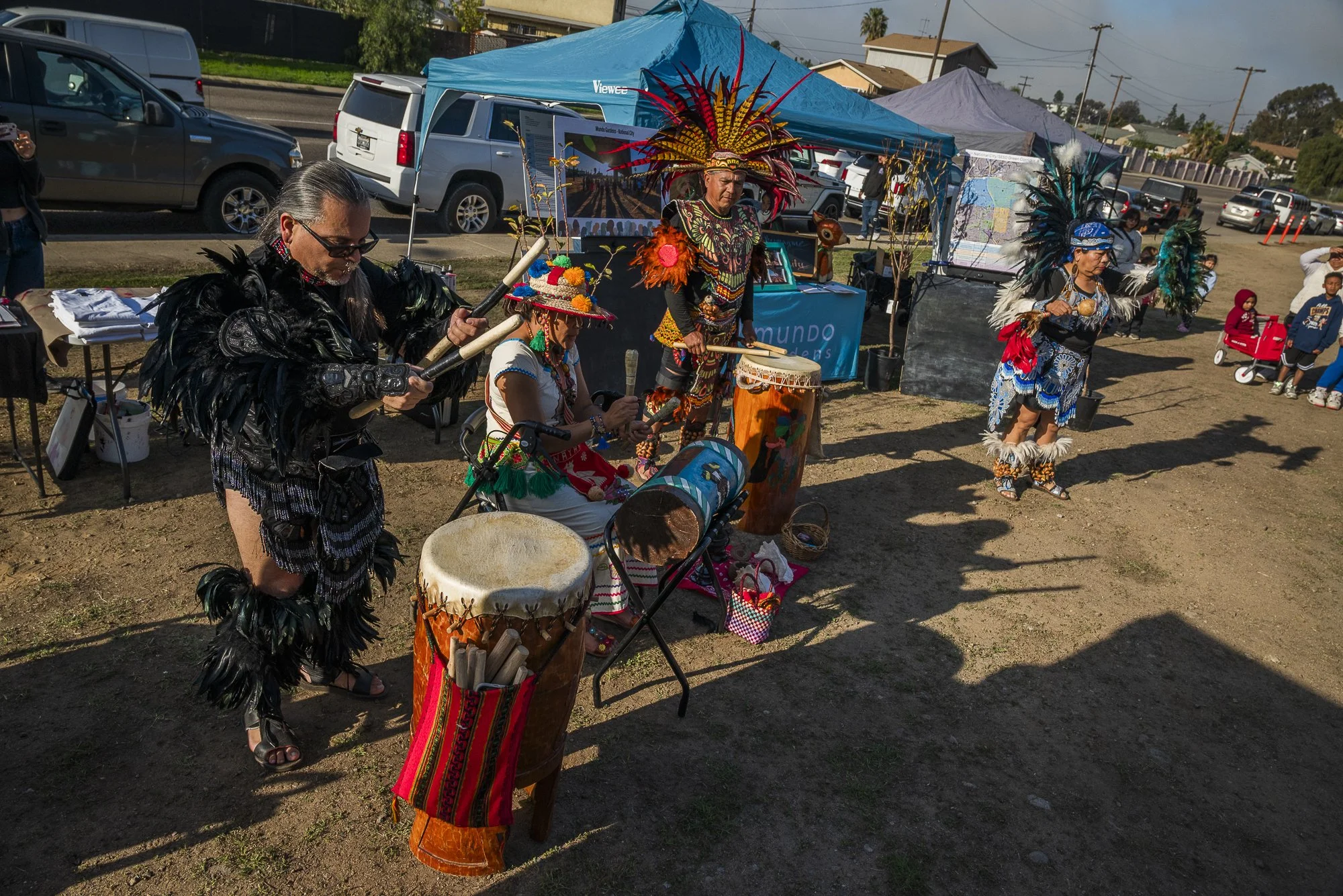 Native American dancers and performers in traditional attire at an outdoor cultural event, with tents and cars in the background.