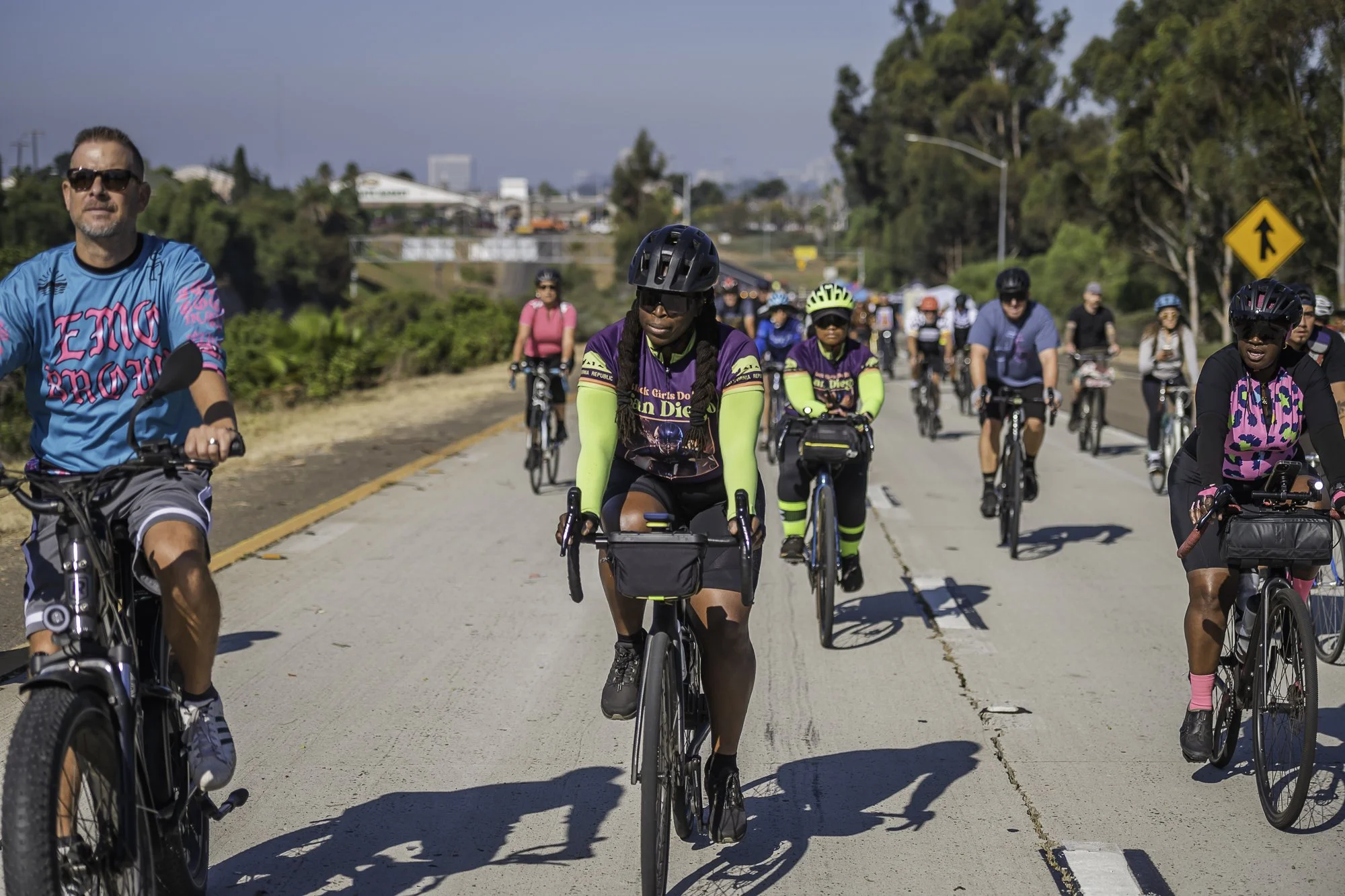 A group of diverse people riding bikes on a sunny day, some wearing helmets, on a dedicated bike lane with trees and buildings in the background.