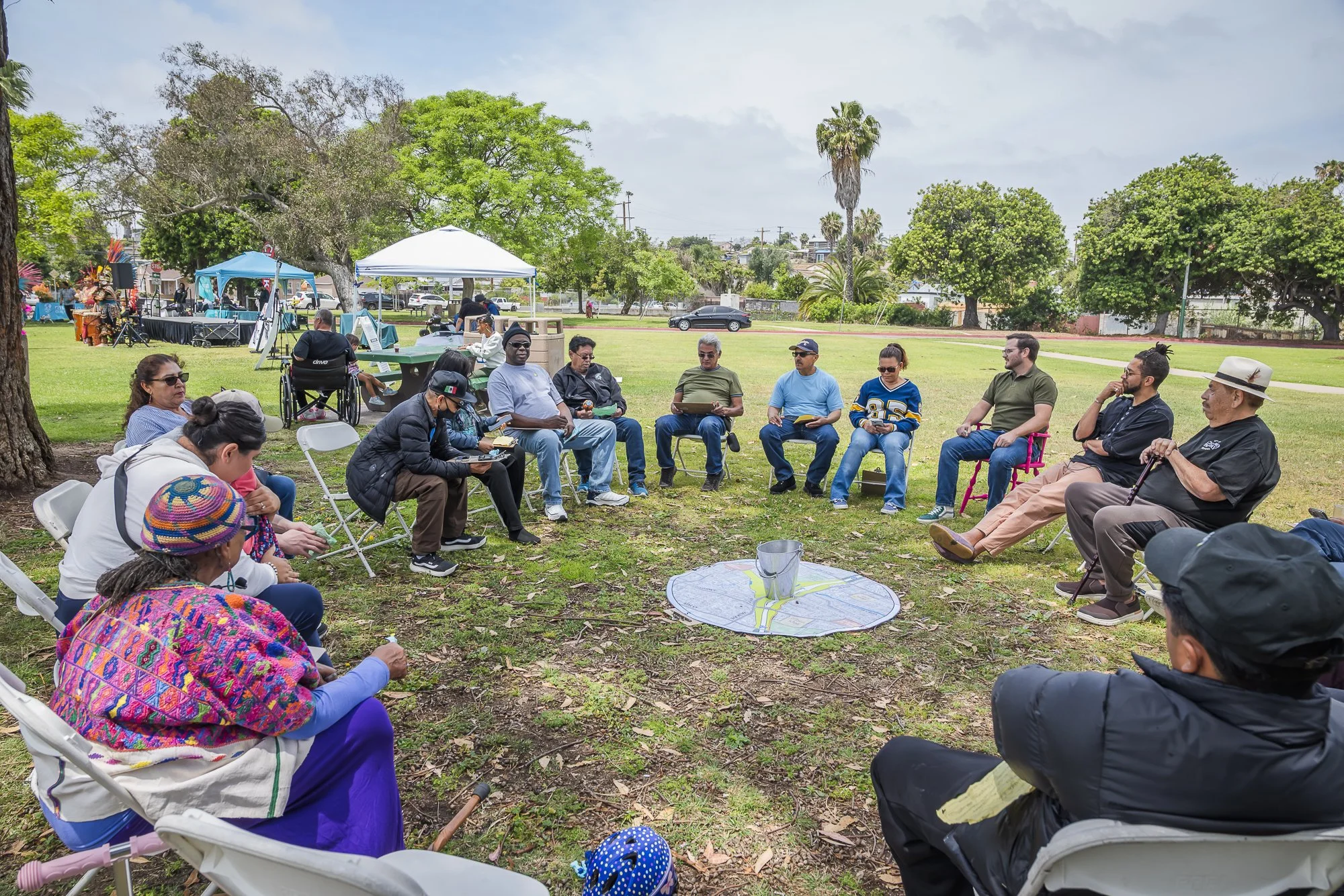 A group of diverse people sitting in a circle in a park during a sunny day, some with notebooks and others listening, with trees, tents, and a few parked cars in the background.