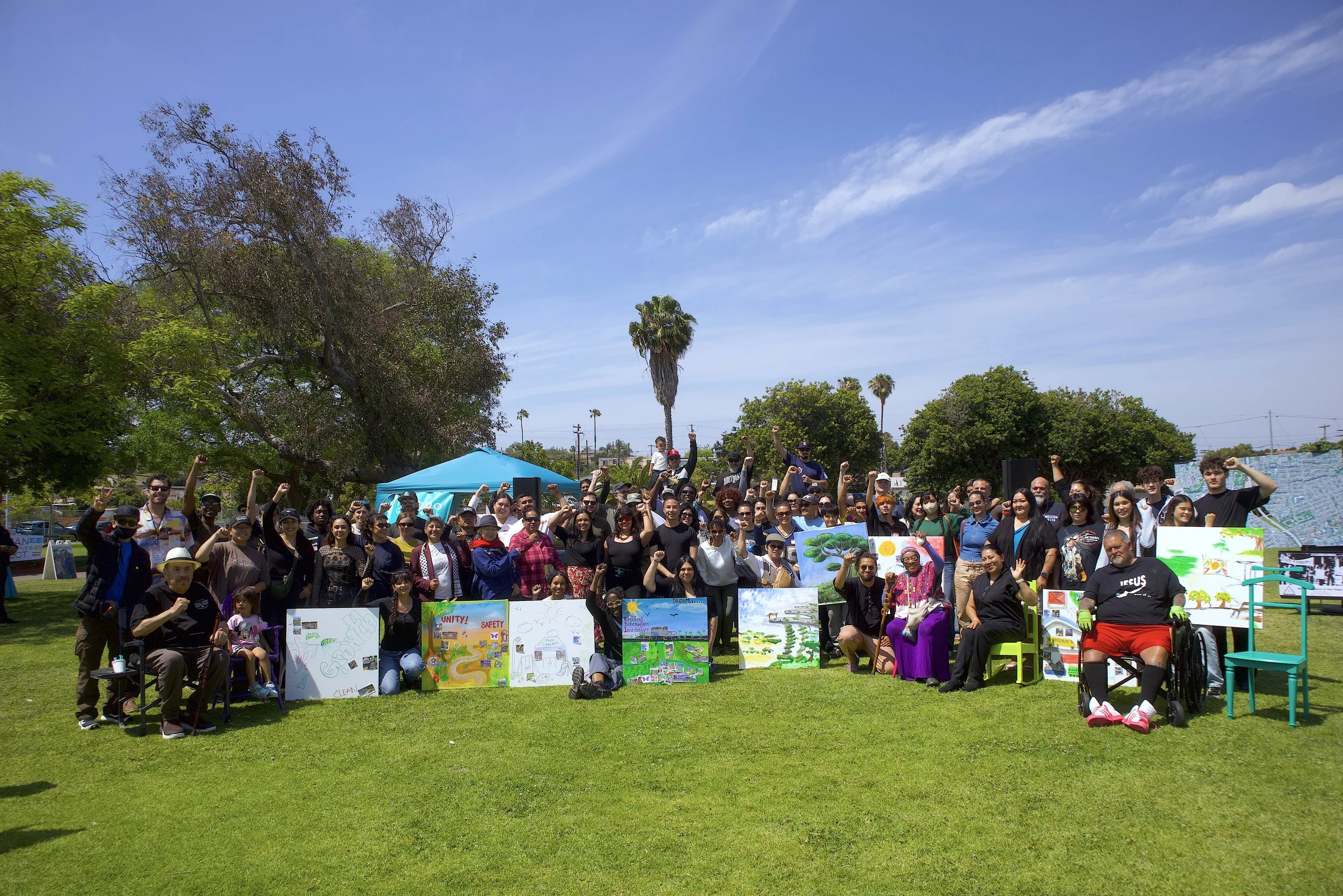 Group of diverse people gathered outdoors at a community event or rally, holding signs and raising fists, with blue sky, palm trees, and green trees in the background.