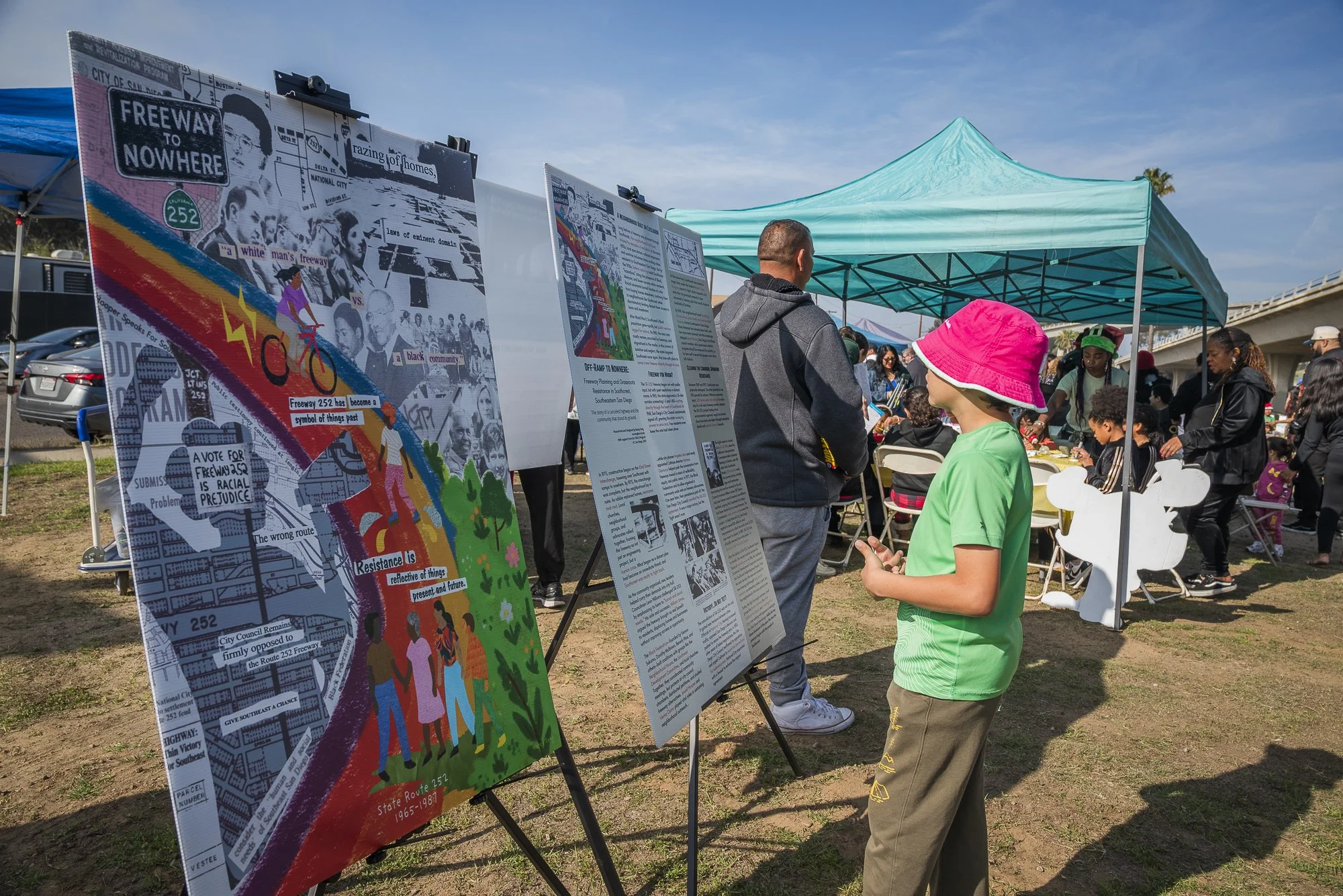 A young boy wearing a bright pink hat and green shirt looking at a large informational poster about freeway 252 at an outdoor event. Several other people are visible, some reading posters, under a blue canopy tent with tables nearby. Vehicles are par