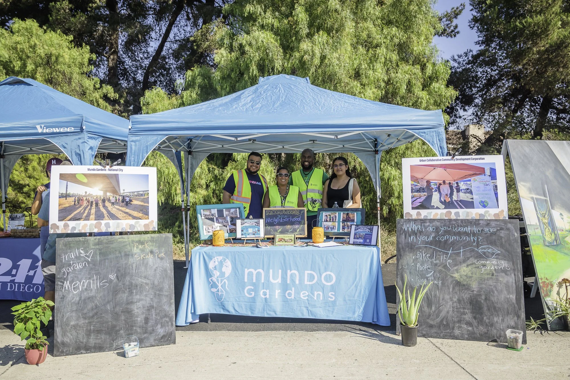 Group of four people standing behind a table at an outdoor community event, under a blue canopy with trees in the background. The table displays informational materials and a sign on the front reads 'mundo garden'. There are two large posters on stan