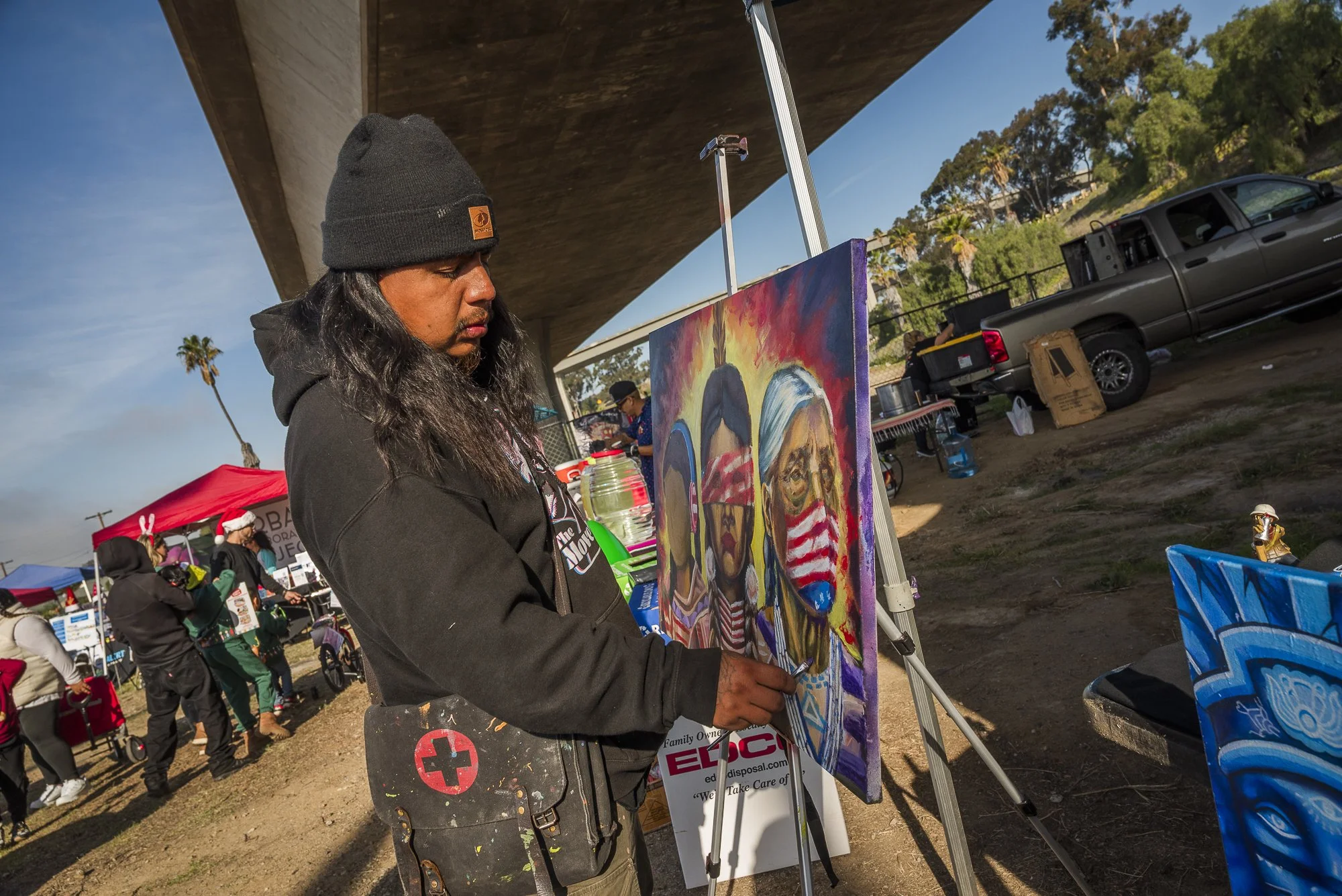 Man with long hair and black beanie wearing a black hoodie, painting at an outdoor art fair. The painting features caricatures of children. Other people and booths are visible in the background under a bridge on a sunny day.