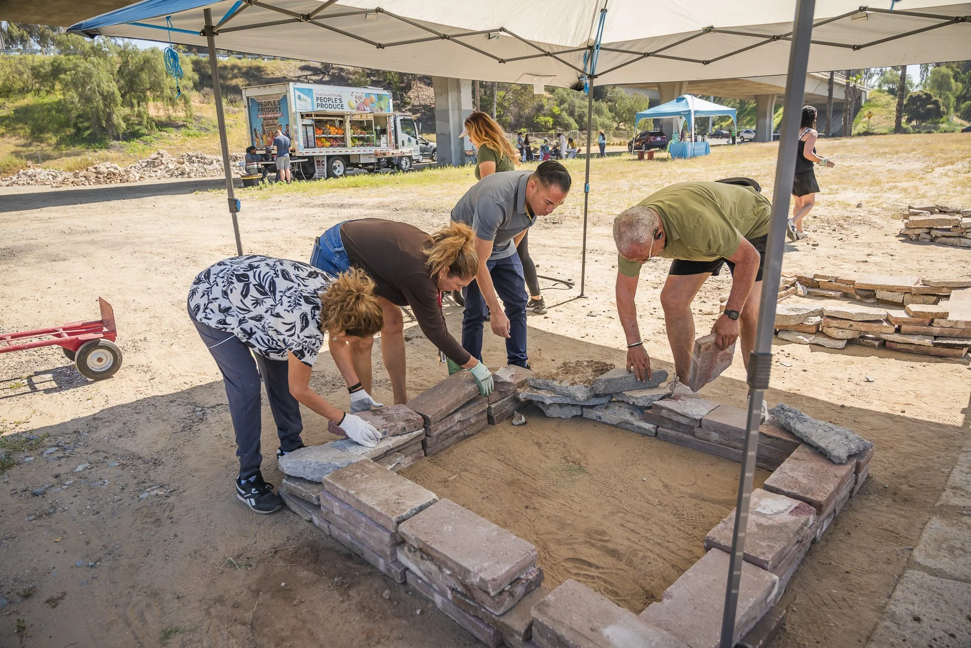 People working together to construct a brick fire pit or garden border outdoors under a canopy, with a food truck and trees in the background.