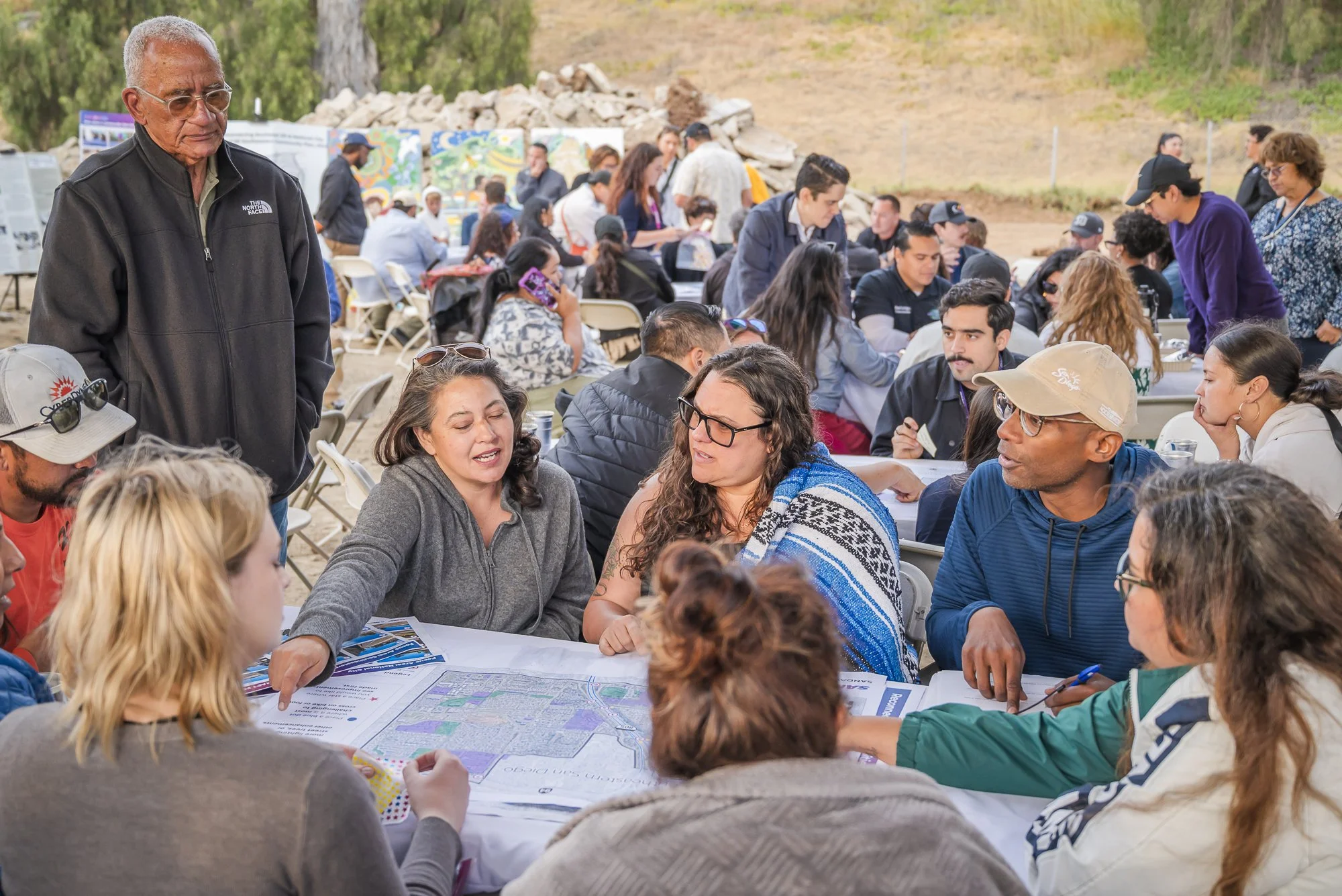 A large group of people gathered outdoors, sitting at tables covered with maps and informational materials, engaging in discussion during what appears to be a community or educational event.