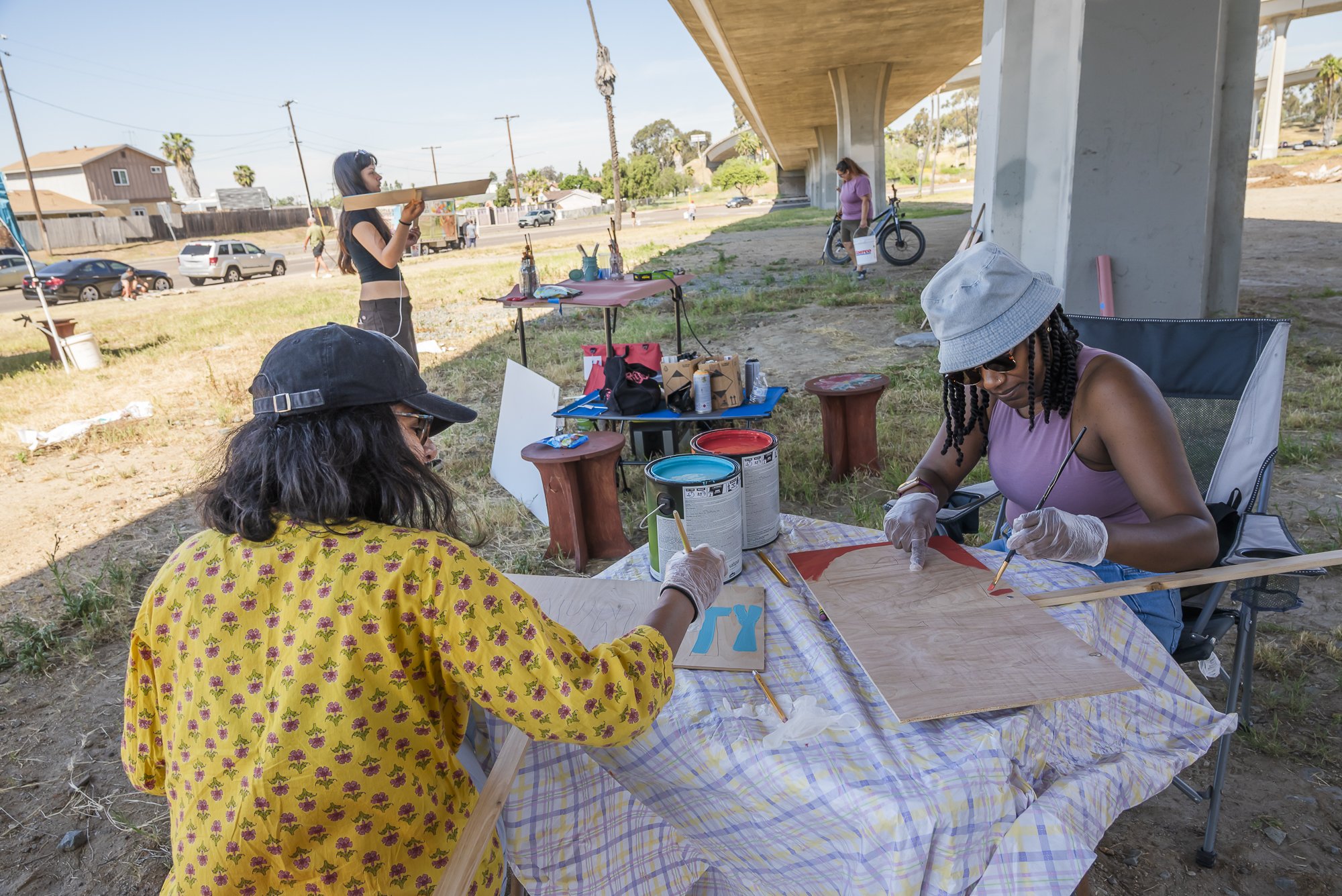 Group of women painting wooden signs outdoors under an overpass on a sunny day.