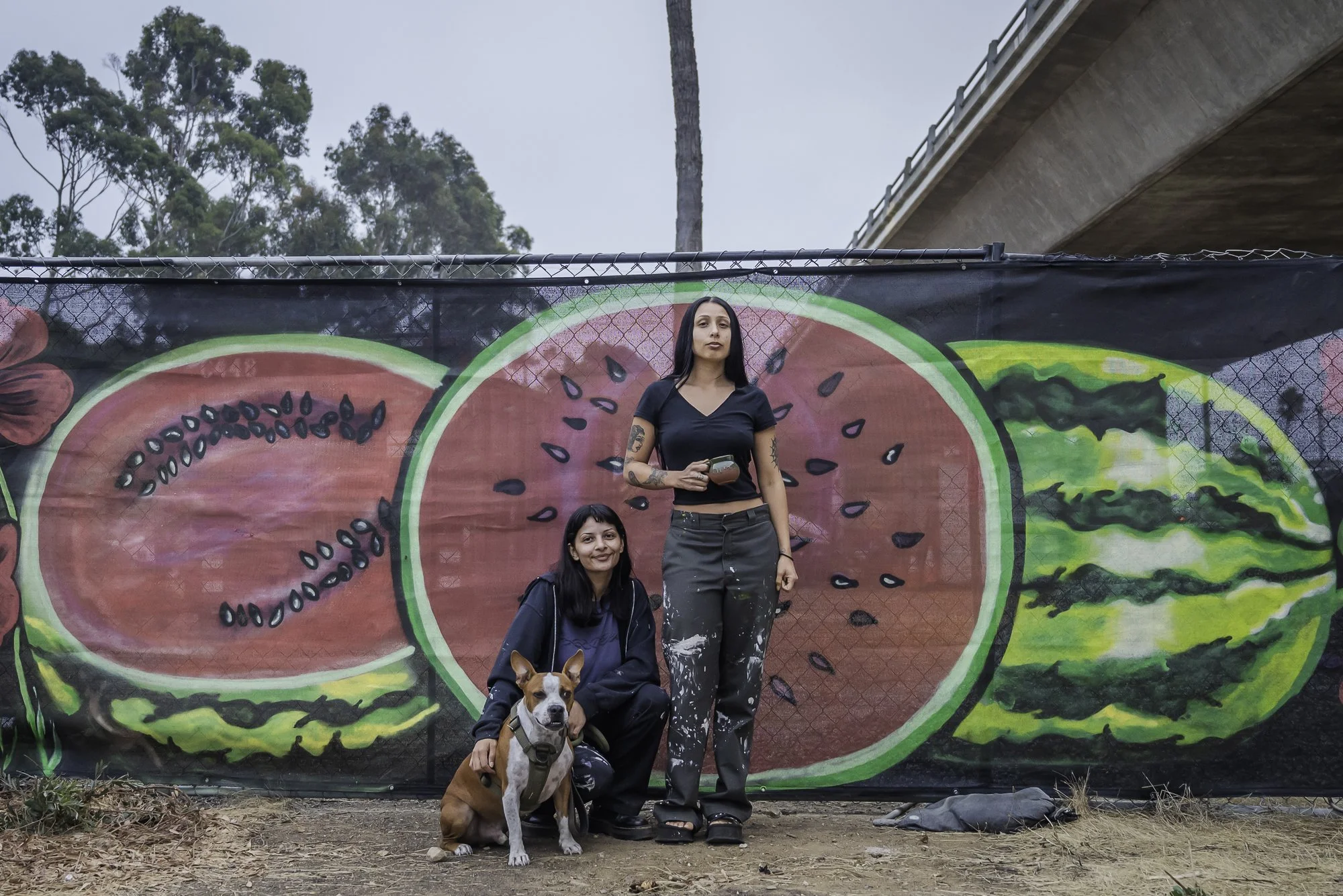 Two women and a dog standing in front of a colorful mural featuring large slices of watermelon and other fruits on a fence at an outdoor location.