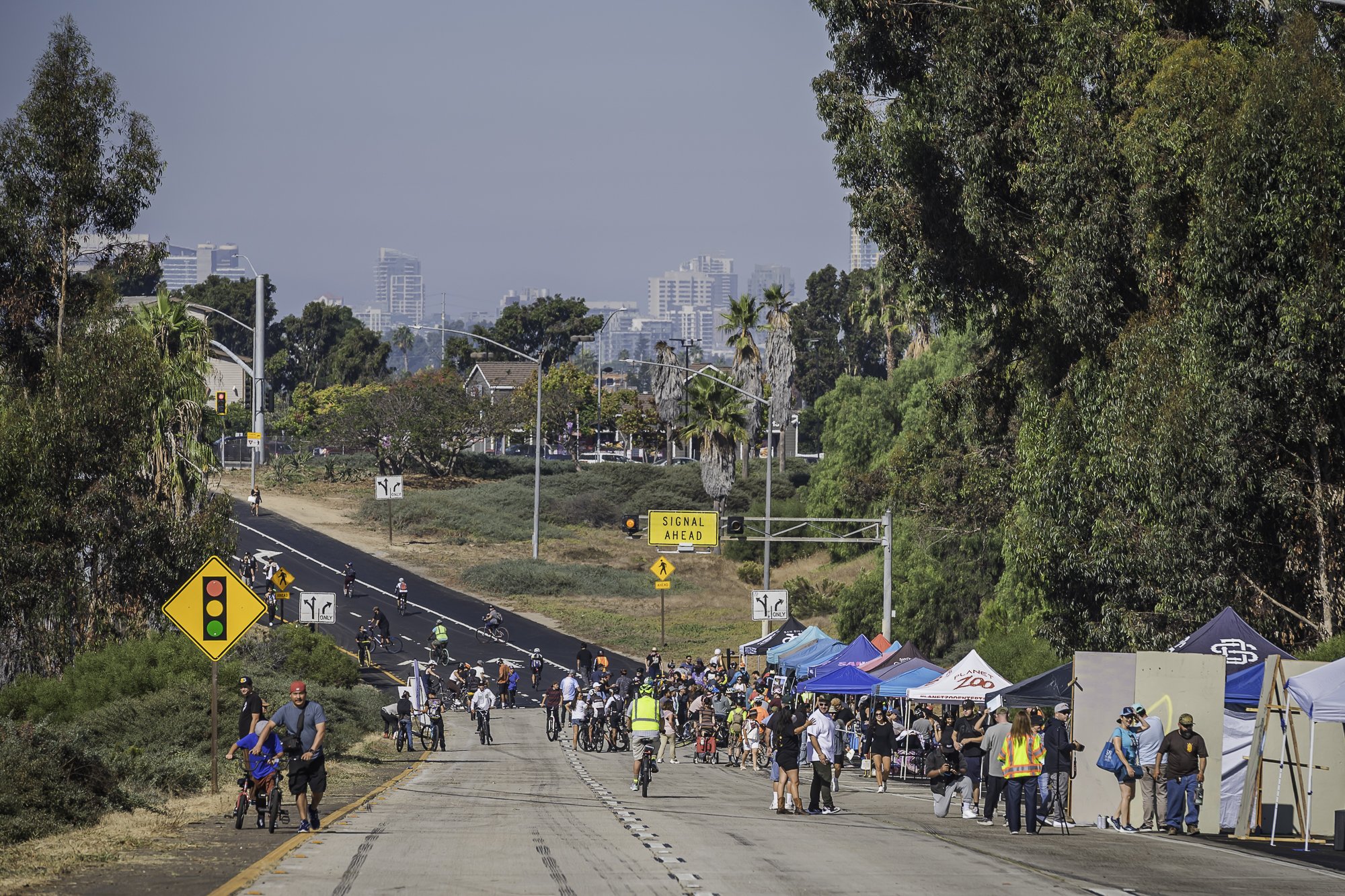 A large outdoor community event with many people, tents, and tents set up along a street with a view of cityscape buildings in the background. Some people are riding bikes, walking, and socializing.