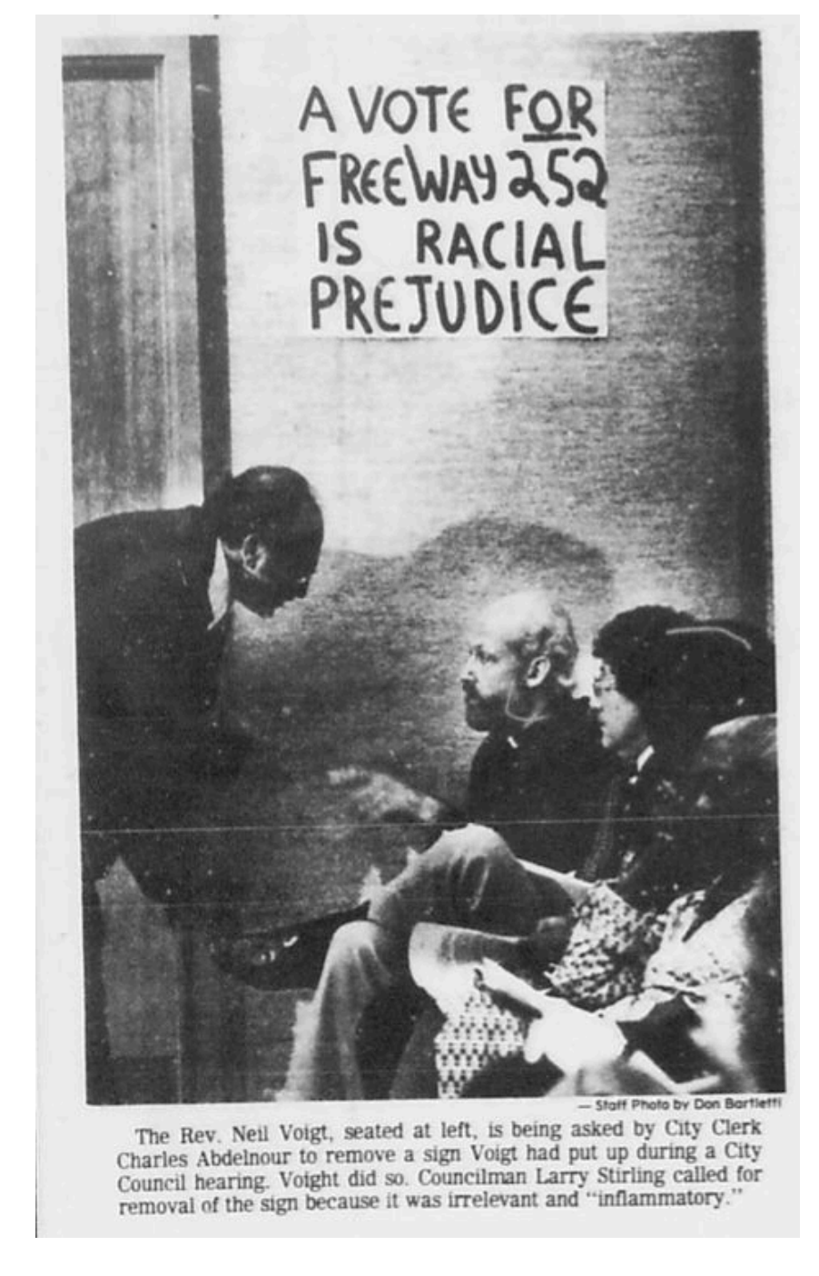 A black-and-white photograph showing a man seated, with a woman sitting next to him, both facing a man standing and leaning forward. Behind them, a sign reads "A VOTE FOR FREEWAY 252 IS RACIAL PREJUDICE." The caption explains that Reverend Neal Voigt was asked to remove the sign during a city hearing, but he refused because he considered the sign irrelevant and inflammatory.
