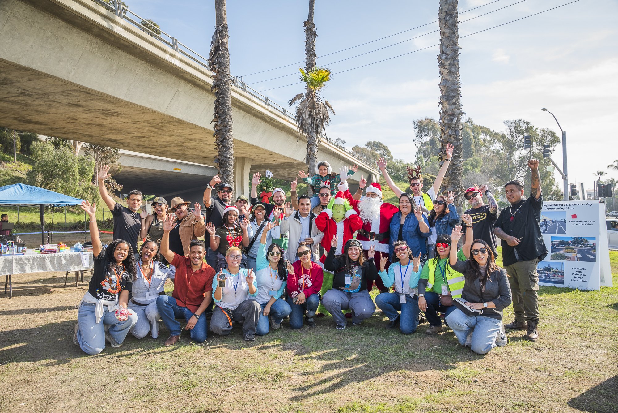 Group of people celebrating outdoors under a bridge, including individuals dressed as Santa Claus and a Christmas elf, with palm trees and traffic signals in the background.