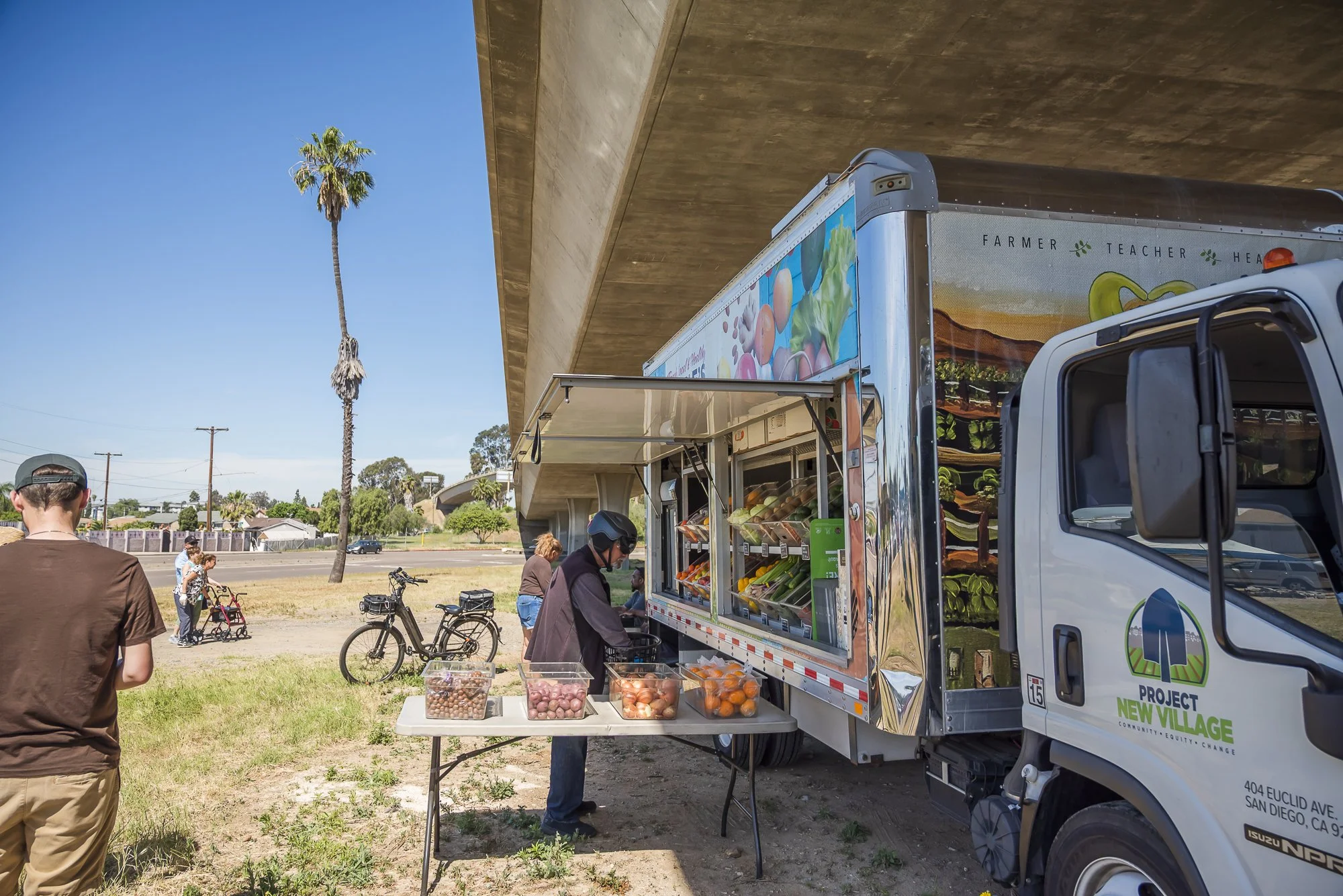A mobile produce stand truck offering fruits and vegetables, with people and bicycles nearby at an outdoor market under a bridge, tall palm trees, and a clear blue sky.