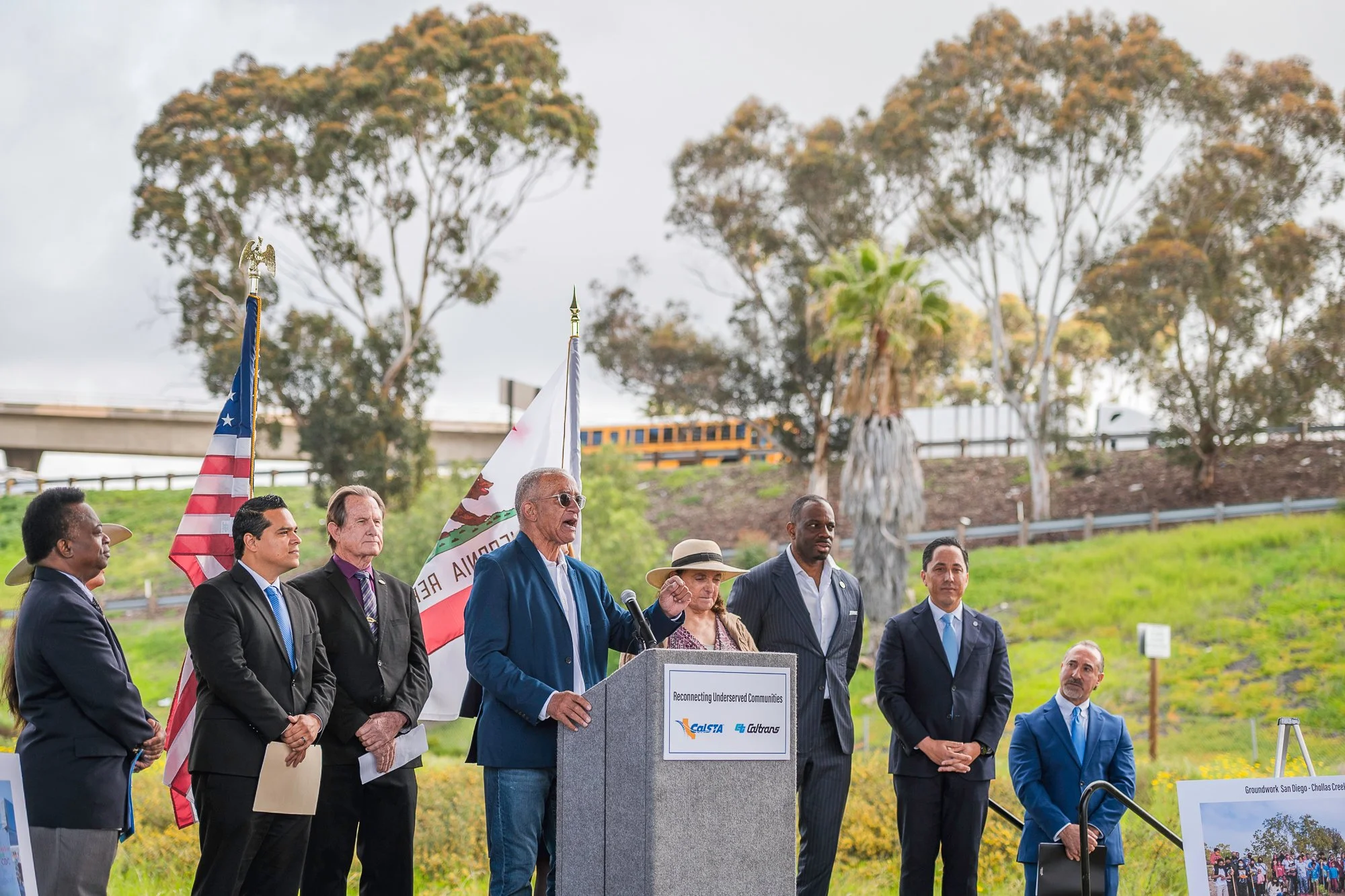A group of diverse people dressed in formal attire standing outdoors during a public event. A man is speaking at a podium that has a sign reading 'Reconnecting Underserved Communities.' Behind them, flags including the California state flag and the American flag are displayed. The background features trees, a highway overpass, and a yellow school bus.