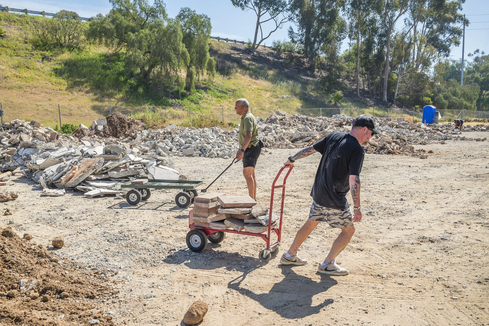 People working on a construction site with debris, rocks, and a portable toilet in the background, some trees on a hill, and a fence.