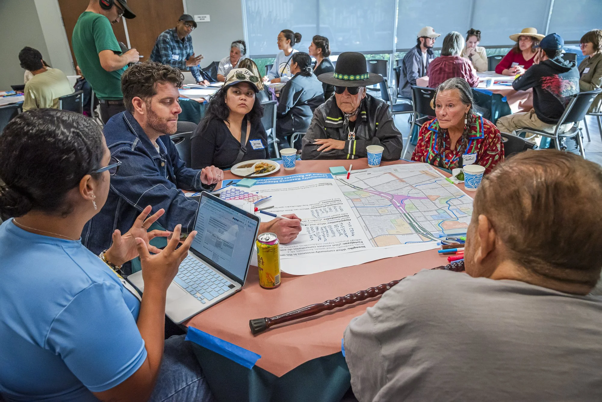 A diverse group of people seated around a large table participating in a meeting or workshop. Some are reading or looking at papers and a laptop, while others listen or engage with each other. The table has maps, notes, and drinks. In the background,