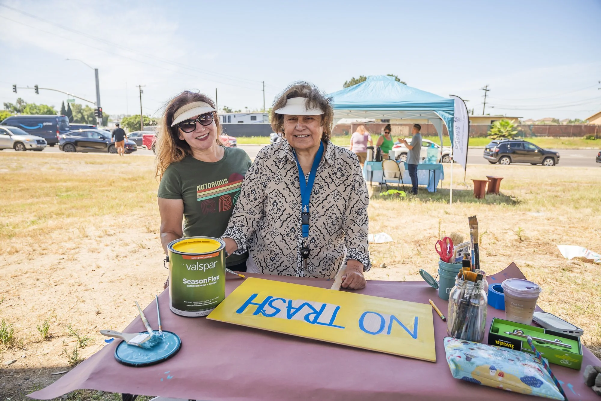 Two women wearing sun visors and sunglasses standing behind a table outdoors with painted signs, art supplies, and a paint can. They are smiling and appear to be volunteering at an outdoor community event on a sunny day.