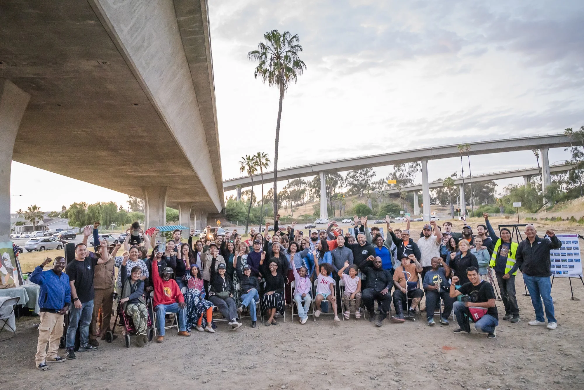 A large group of diverse people gathered outdoors under a bridge, waving and smiling at the camera during a community event, with some children and adults present, and palm trees and cars in the background.