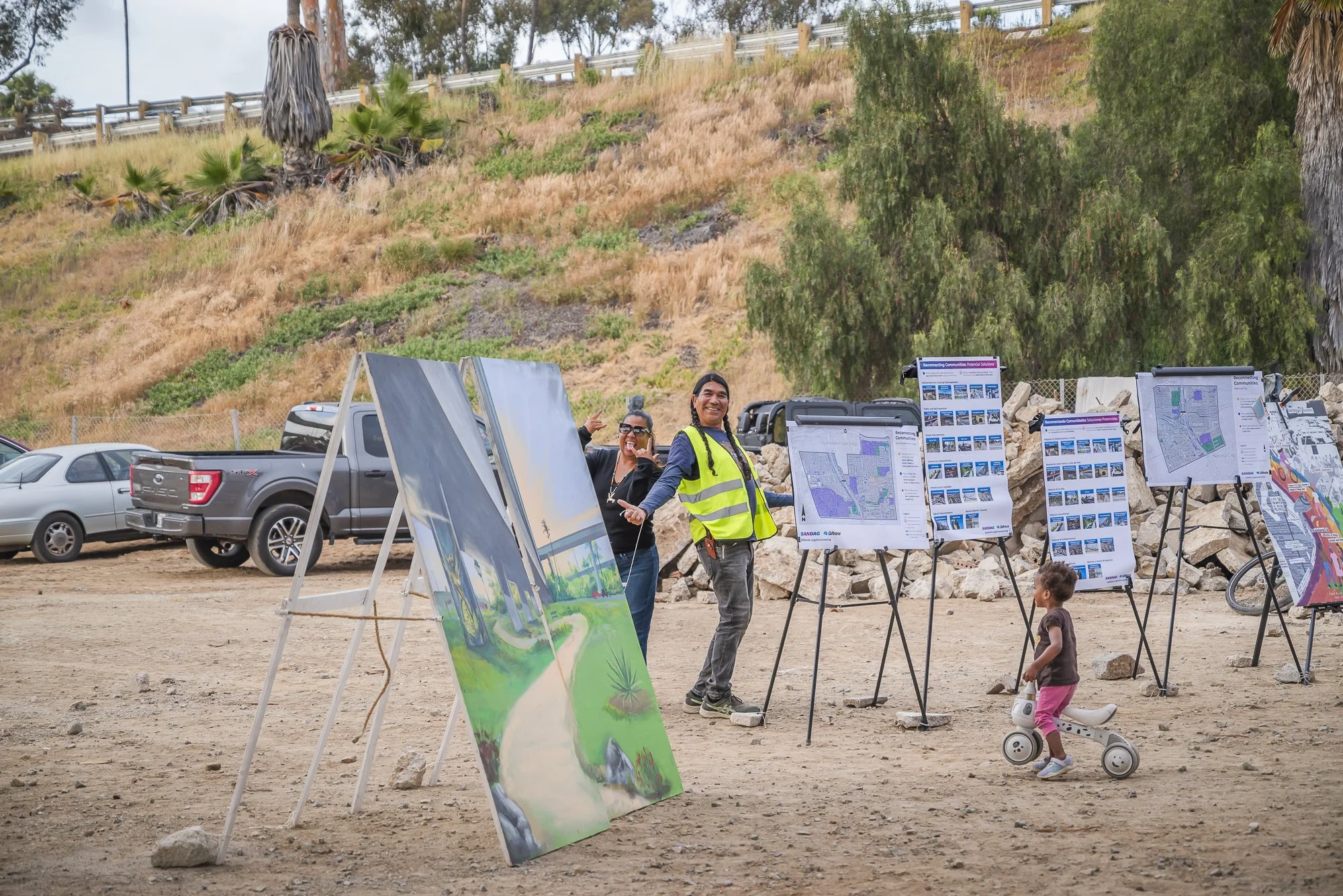 People at an outdoor event with artwork and informational posters on display. A woman in a black top and sunglasses is smiling and talking to a man in a yellow safety vest, who is pointing to an artwork on an easel. A young girl on a scooter is nearb