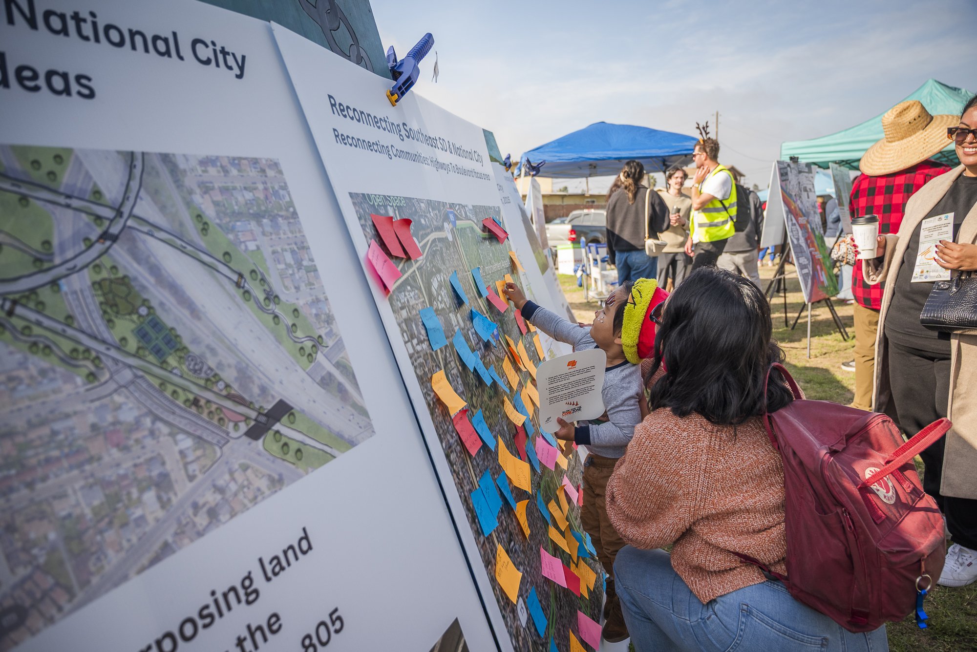 People at an outdoor event gathered around a large informational display board, which features maps and colorful sticky notes, and are engaging in discussions.