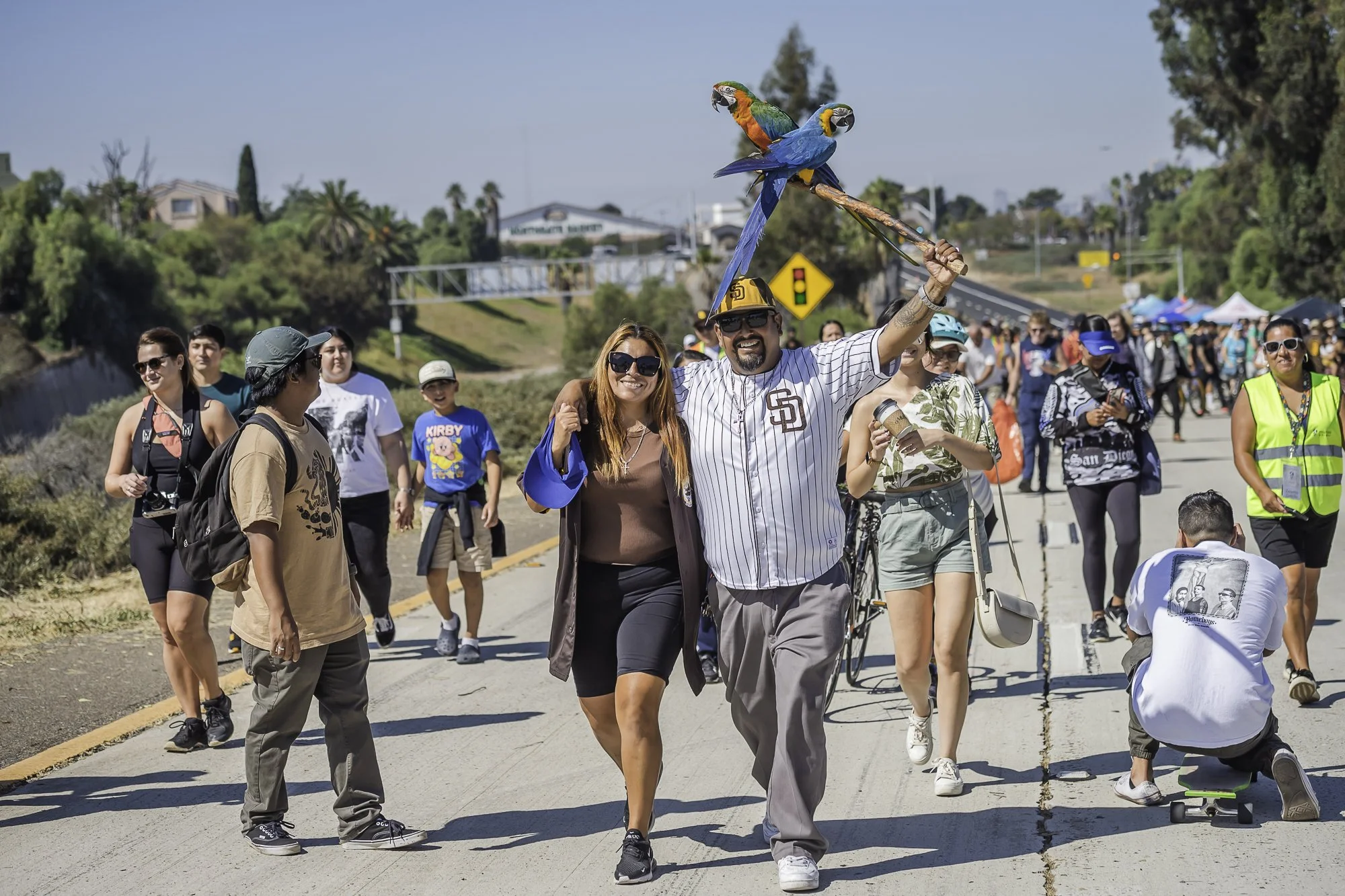 A group of people walking in an outdoor event, with some wearing casual clothing and others in athletic wear. One man is holding two colorful parrots, a macaw and a blue-and-yellow parrot, on his hand. The background shows trees, buildings, a traffic