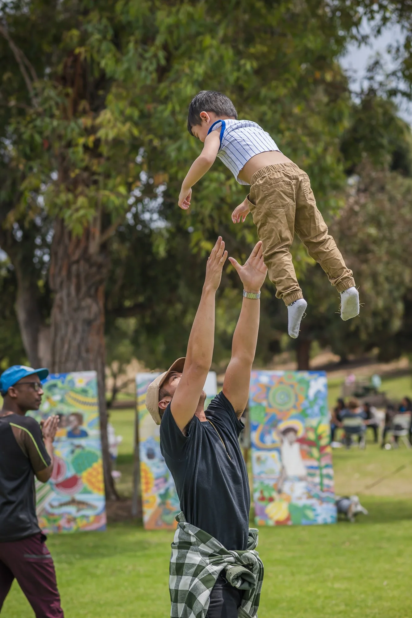 A man wearing a hat lifts a young boy into the air during an outdoor event with colorful artwork and other people in the background.