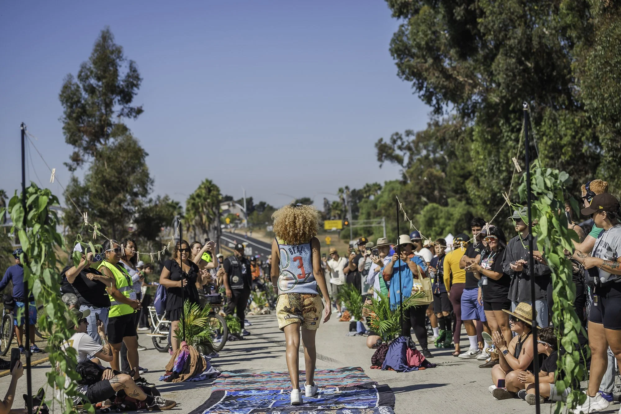 A diverse group of people gather along a sidewalk to watch a person walking in a street festival with a clear blue sky and trees in the background.