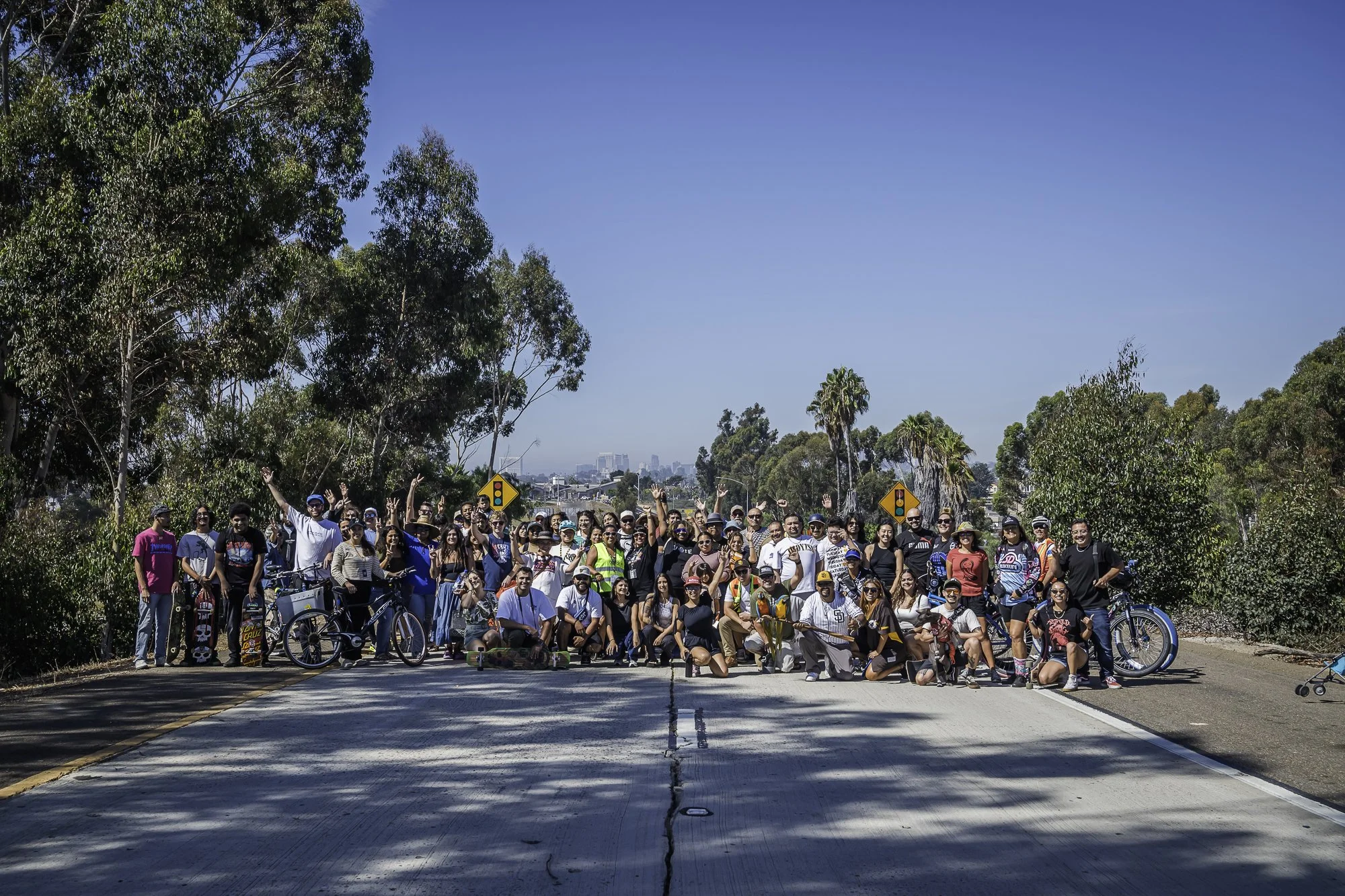 A large group of diverse people gathered together outdoors on a sunny day, some holding bicycles and skateboards, with trees and a clear blue sky in the background.