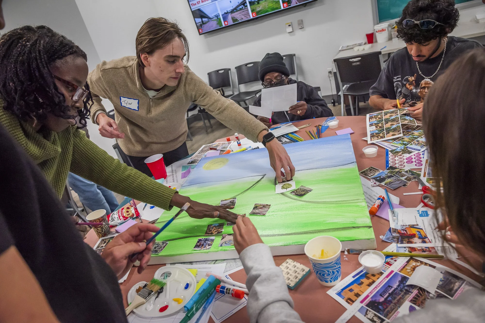 A group of diverse people working on a large landscape painting at a table filled with art supplies, photographs, and printed images, with a monitor displaying images in the background.
