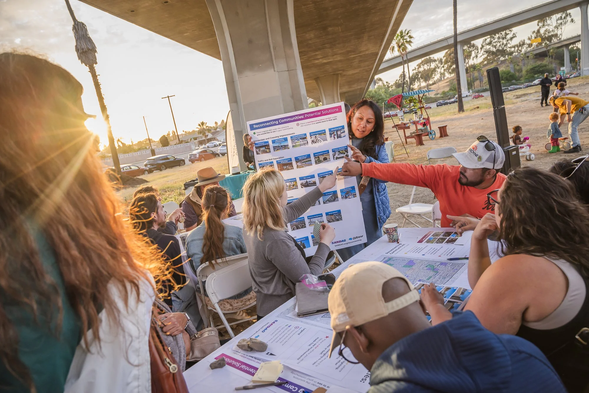 A community meeting taking place under a bridge in the late afternoon sunset. A woman is presenting a large visual display about potential solutions for reconnecting communities. Several elderly and young residents are seated at the tables, some with