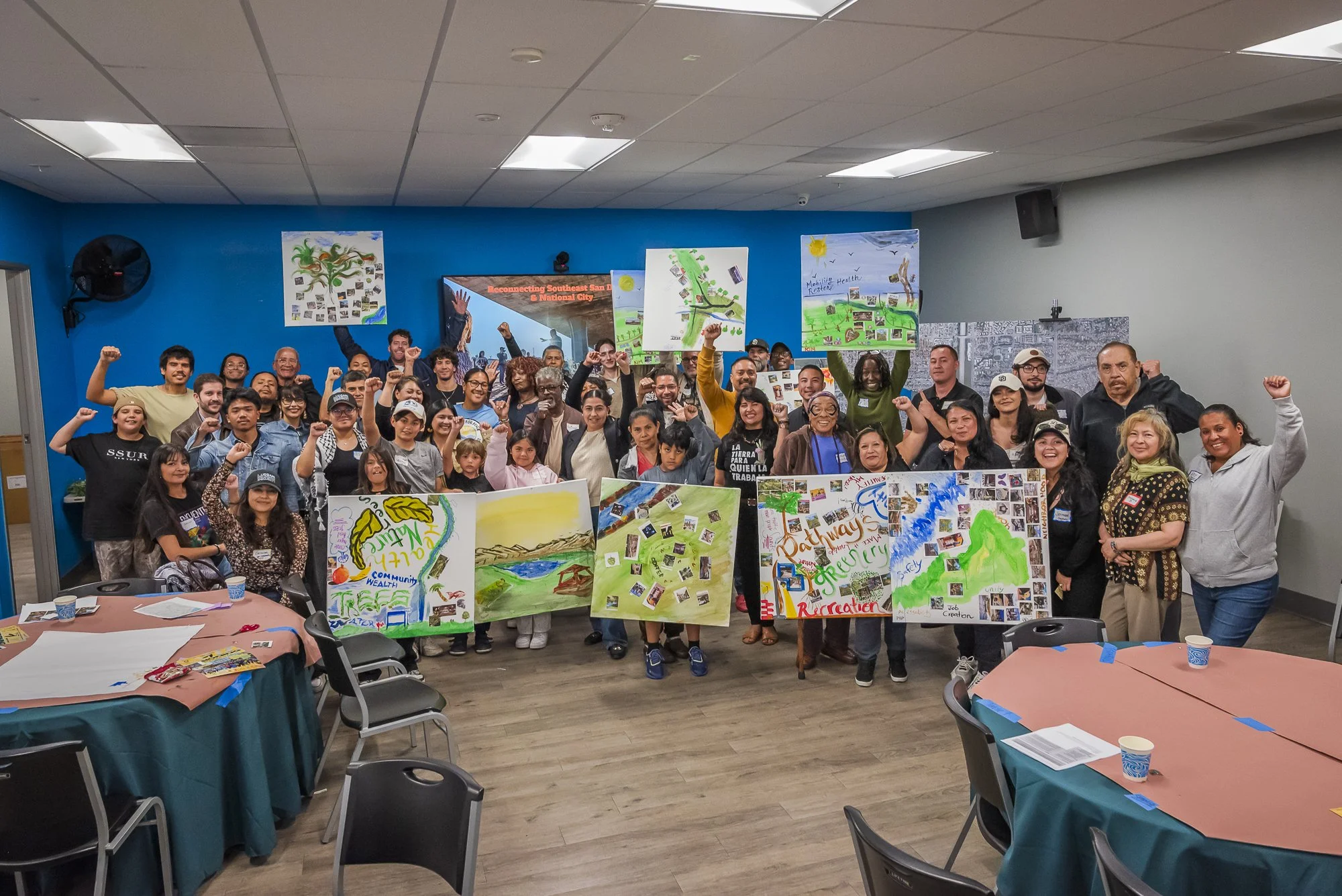Large group of people at an indoor event, holding colorful posters related to community, nature, and recreation, with tables and chairs in the foreground.