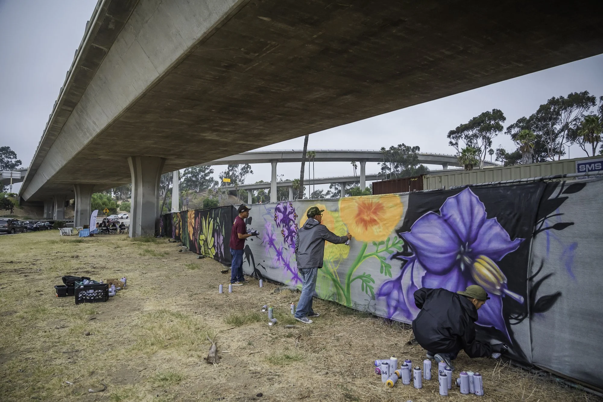 People painting a large colorful mural of flowers on a fence under an overpass. Spray paint cans are scattered on the ground nearby.