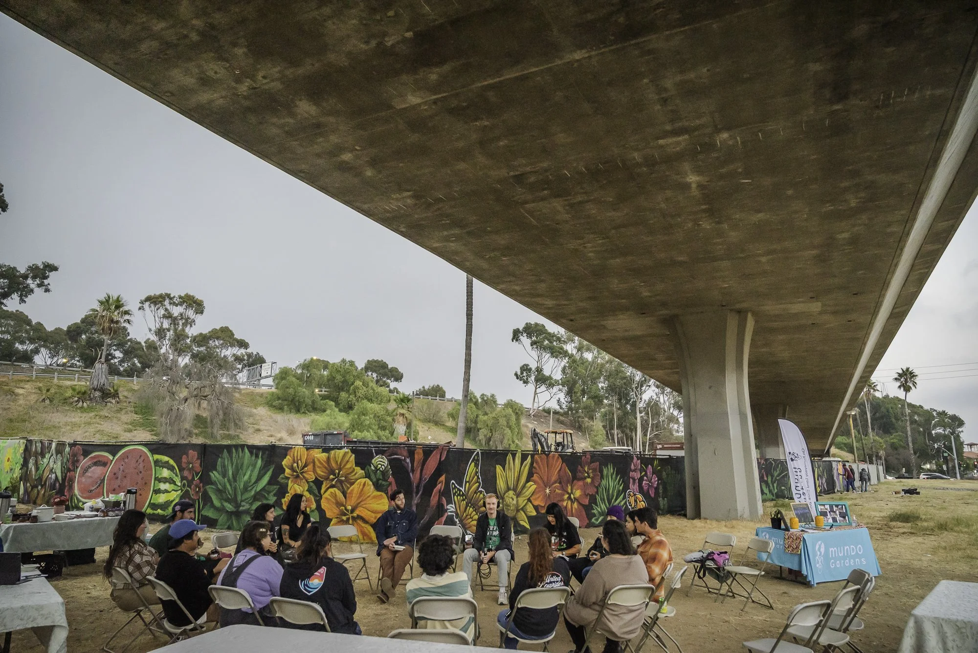 People sitting in a circle outdoors under a large concrete overpass, engaging in a group discussion or workshop, with graffitied wall and greenery in the background.
