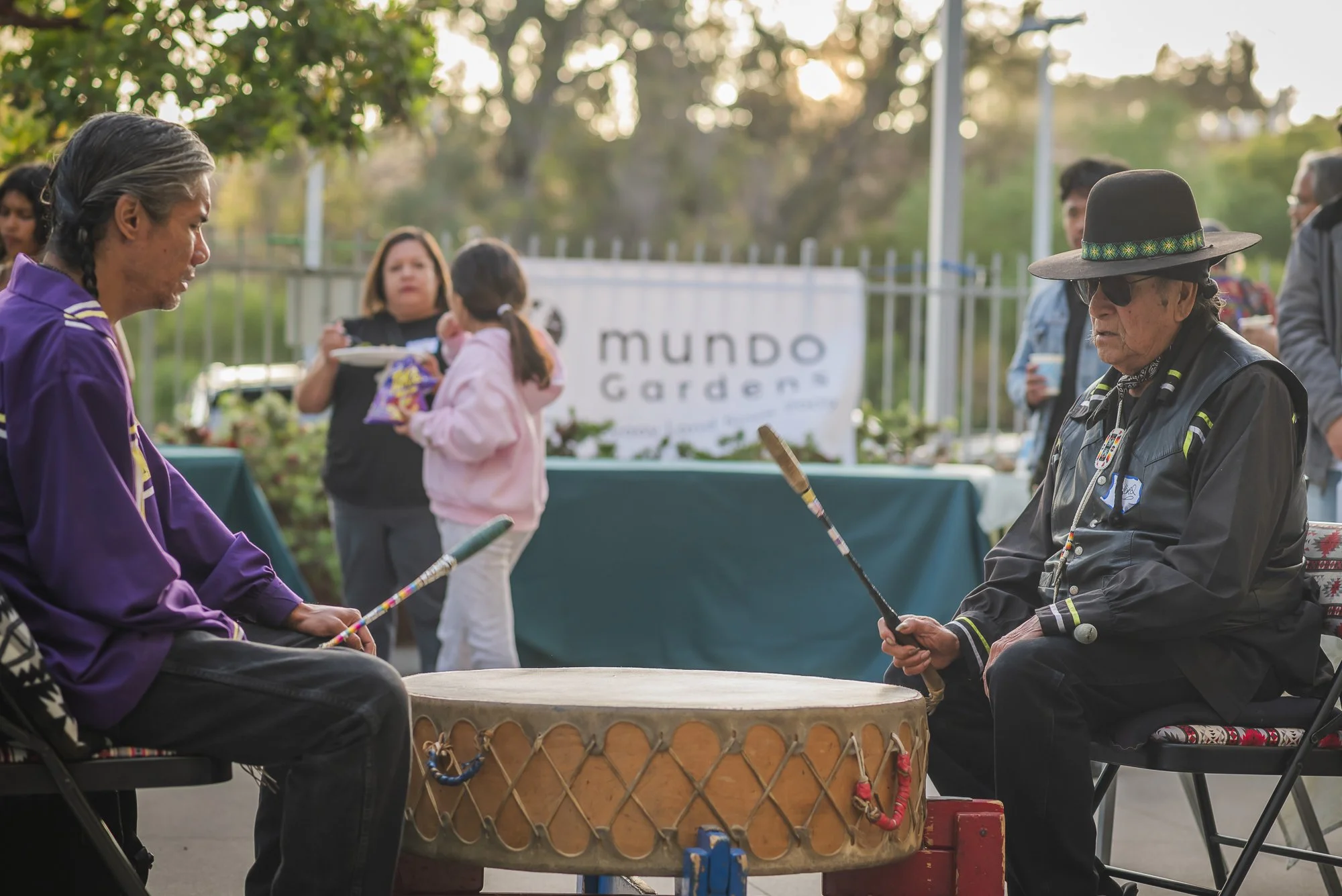 Two Indigenous men sitting around a traditional drum, with people in the background at Mundo Gardens, an outdoor community event.