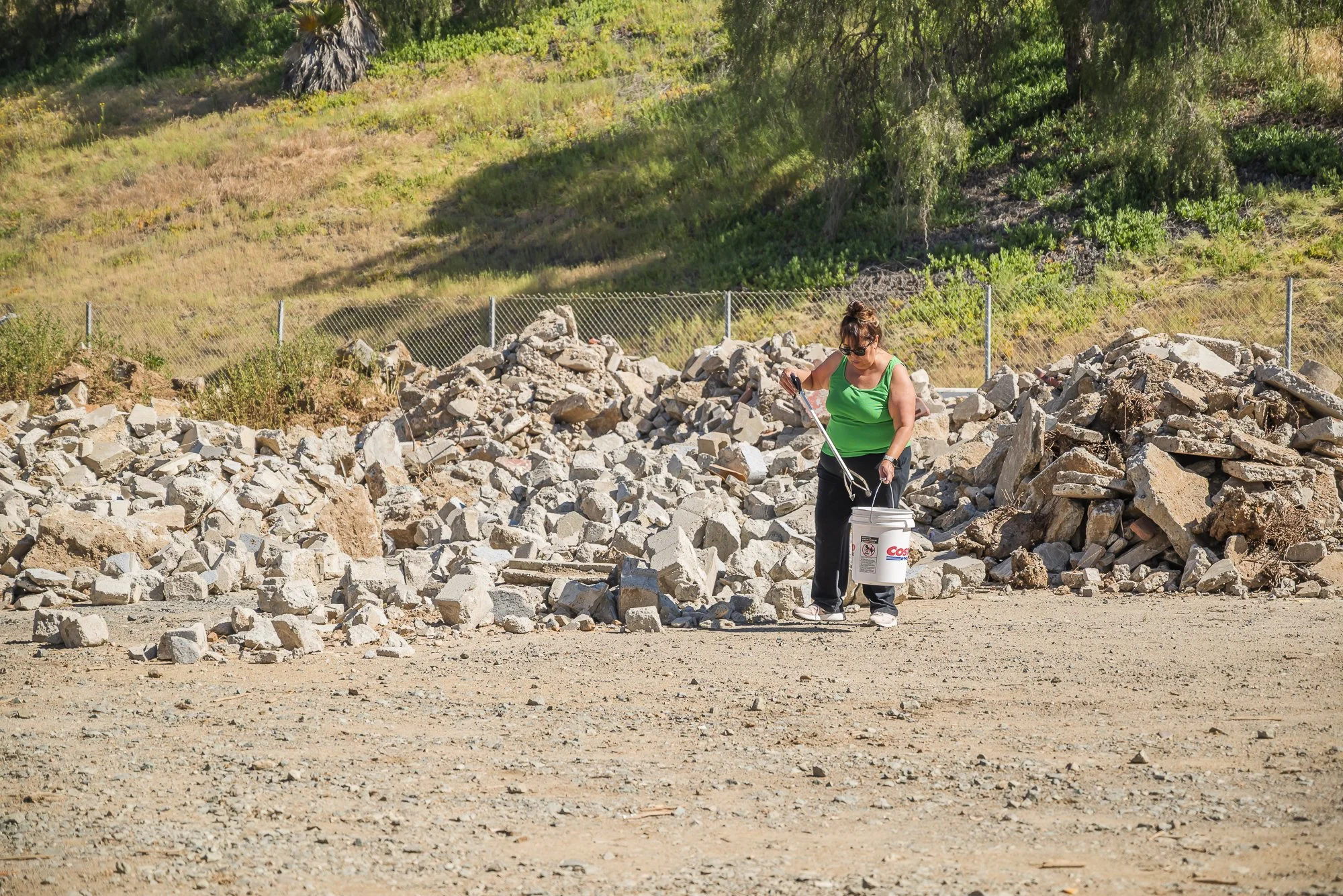 A woman in a green top and black pants holds a bucket and a tool while standing among a pile of rocks at a construction or demolition site, with greenery and trees in the background.