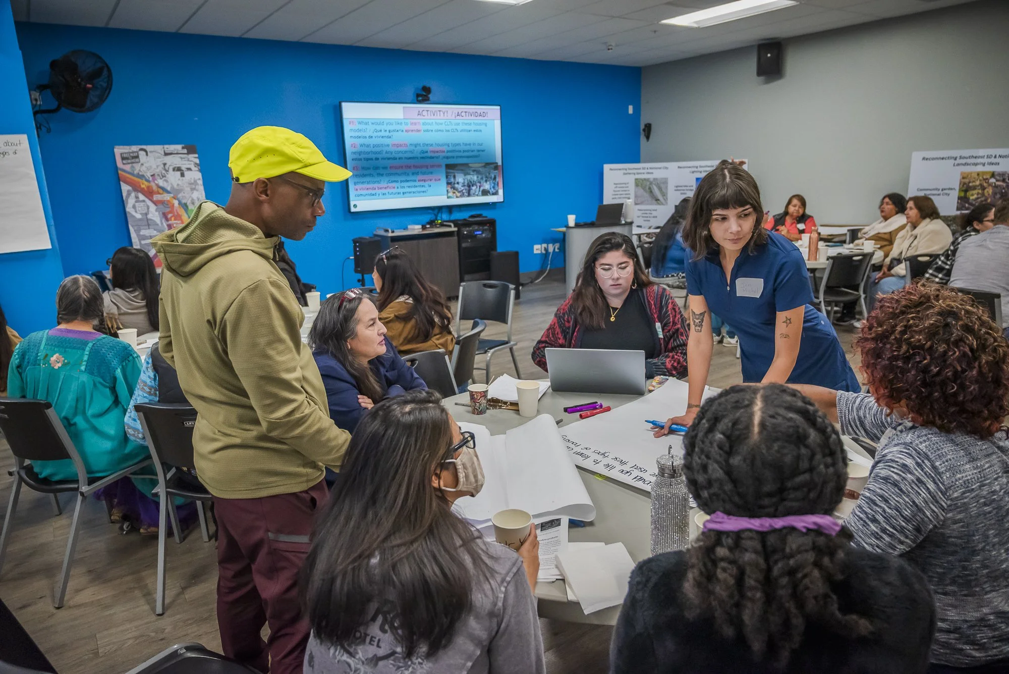 A diverse group of people participating in a workshop or discussion in a conference room. Some are seated around tables, others are standing and engaging with documents and devices. In the background, a large screen displays activity instructions and