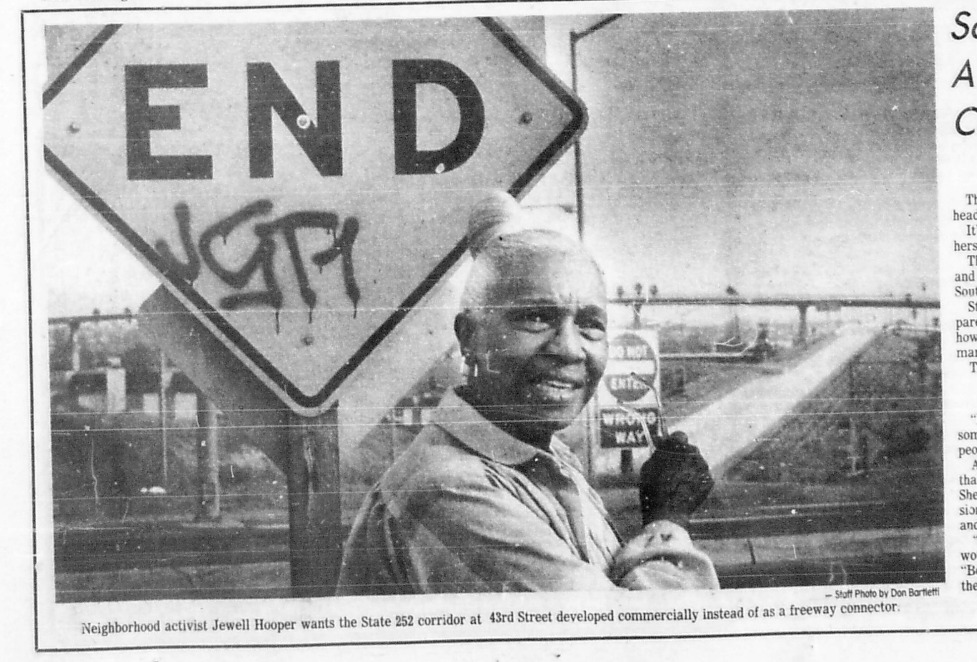 A black and white photograph of a smiling woman holding a sign that reads 'END' in large block letters. The photo appears to be taken at a street event with traffic in the background.