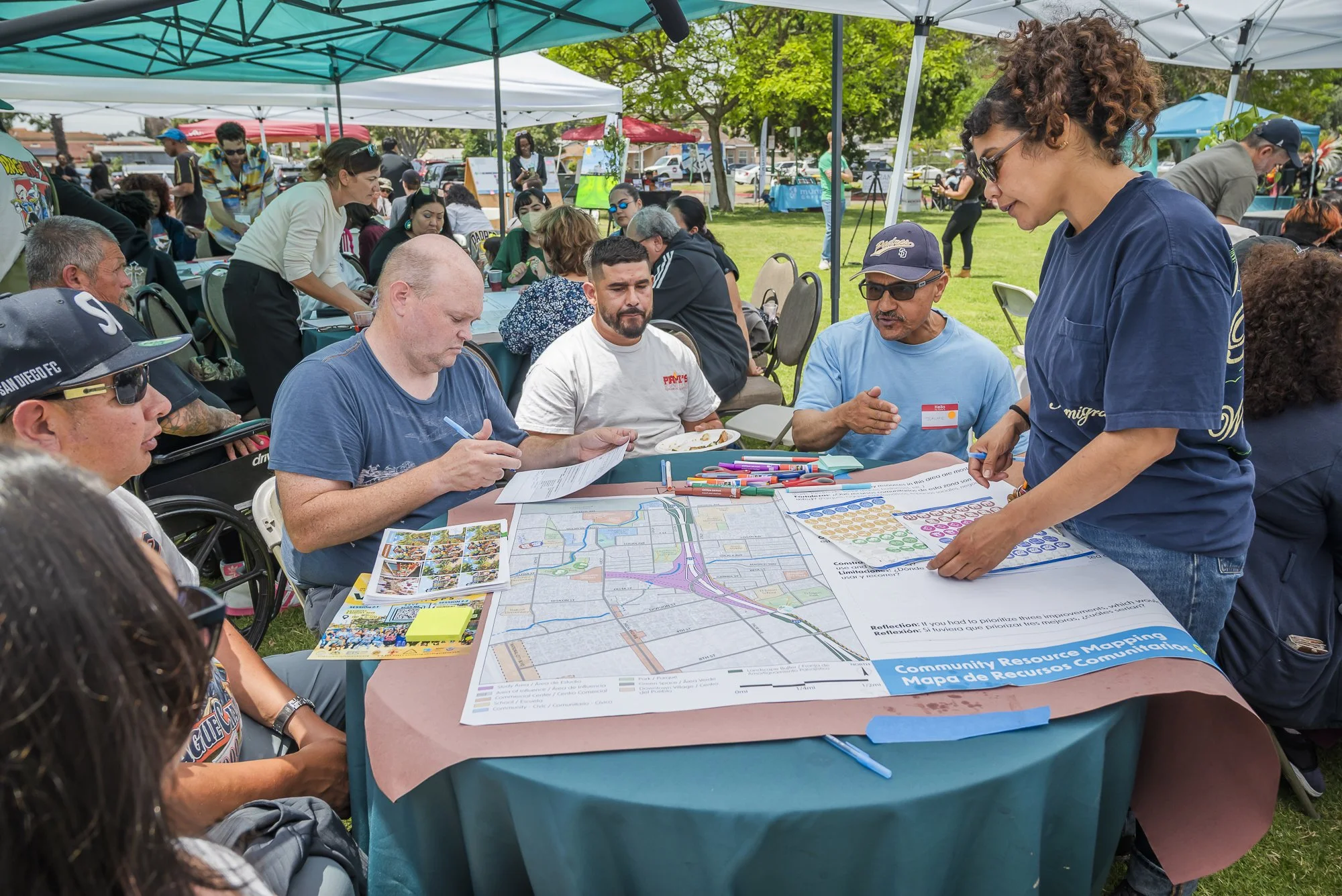 People sitting at a table outdoors under tents, engaged in discussion, with maps and materials spread out on the table during a community event.