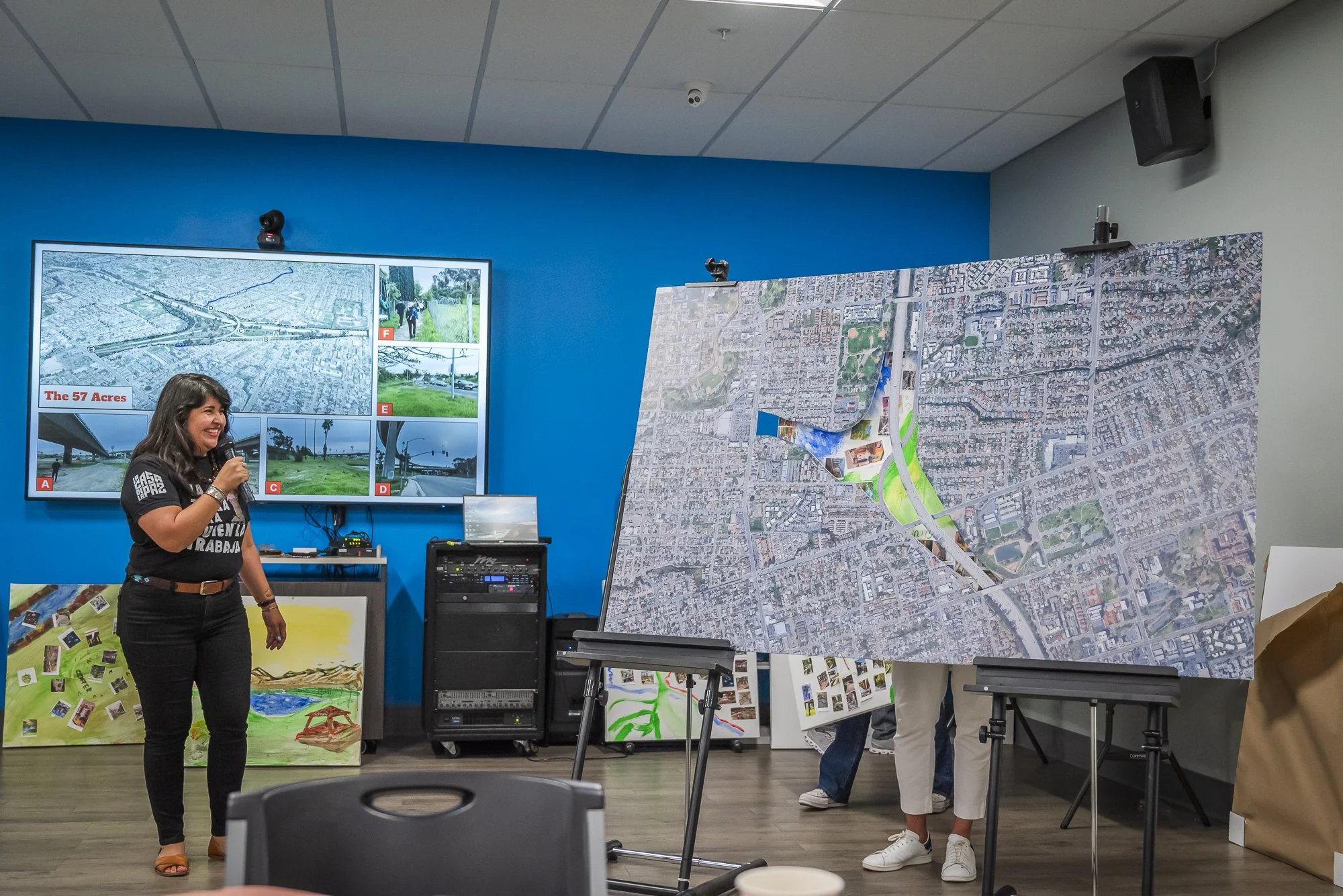 A woman with dark hair holds a microphone while speaking in front of a large displayed map. There are other people partially visible behind the map, and the room has blue and gray walls with ceiling tiles. Various artwork and electronic equipment are
