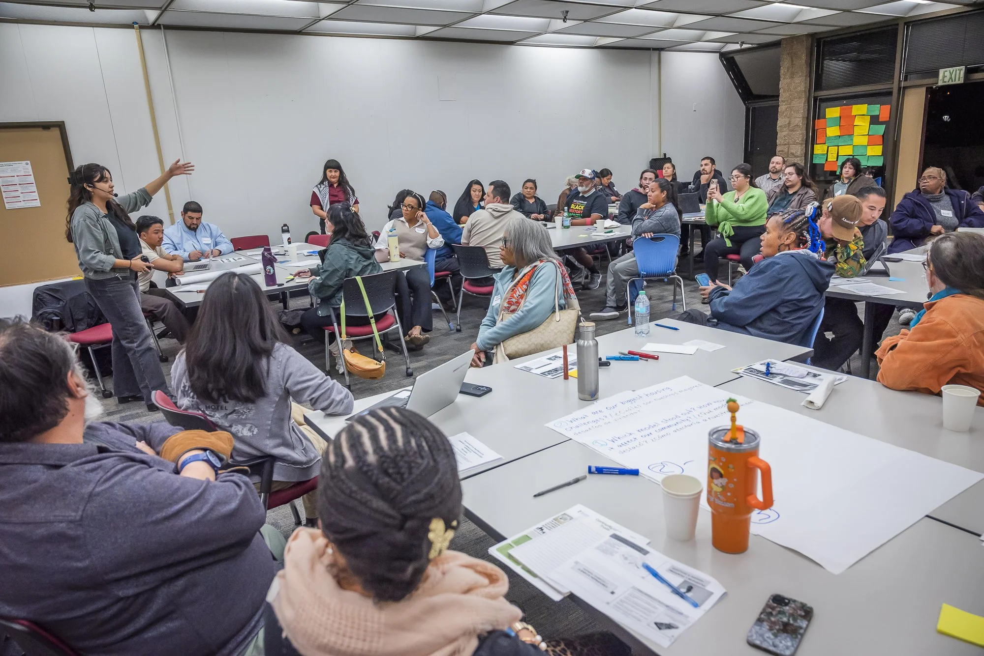 Group of people attending a workshop or seminar in a conference room, some taking notes, listening to a woman presenting at the front.