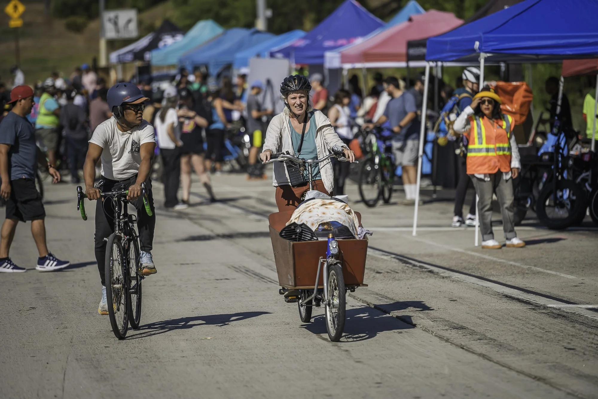 People at an outdoor market or festival, with tents set up, including a woman riding a bike with a baby in a cargo attachment, and others walking and browsing tents.