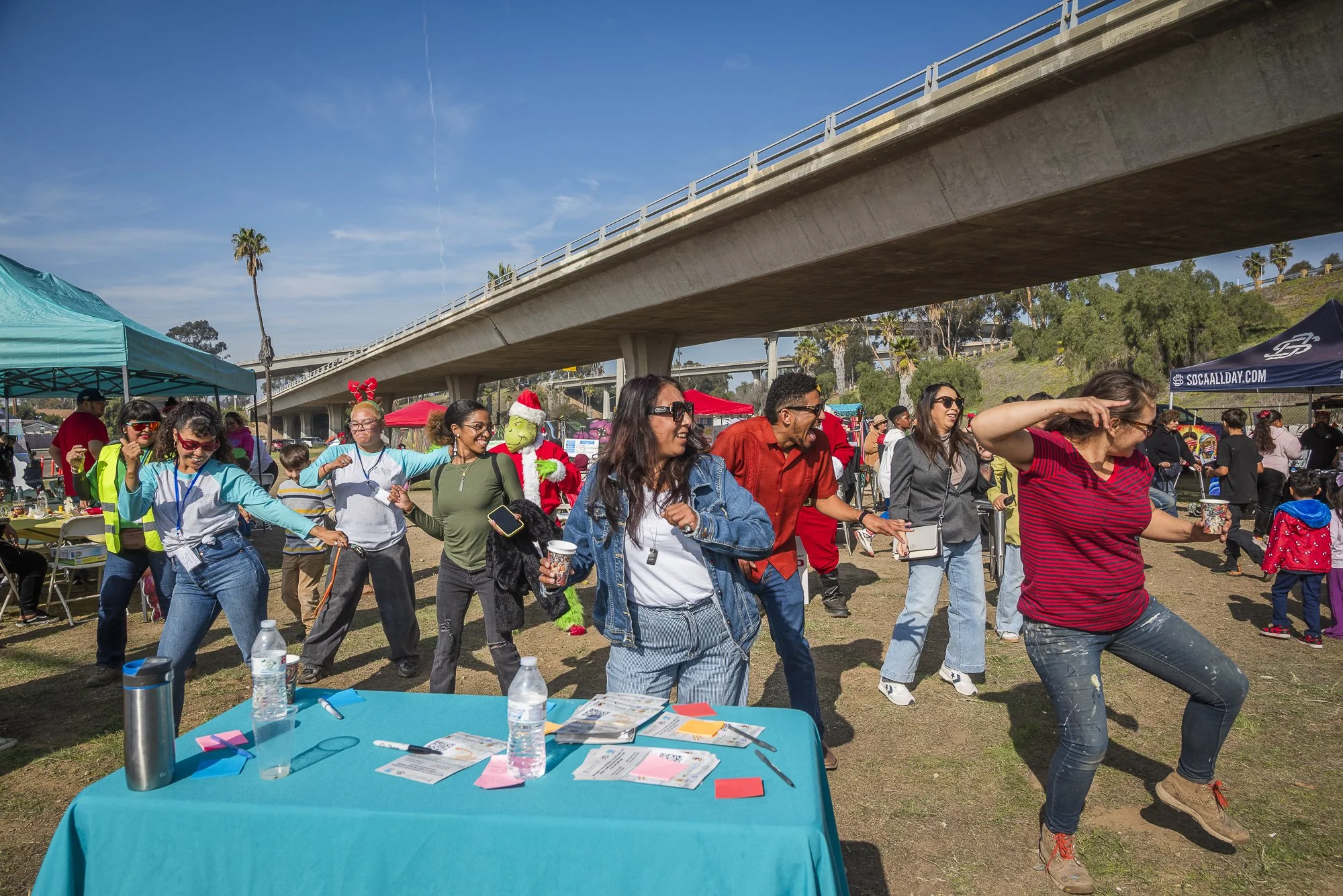 People gathering and dancing outdoors under a bridge during a festive event on a sunny day.