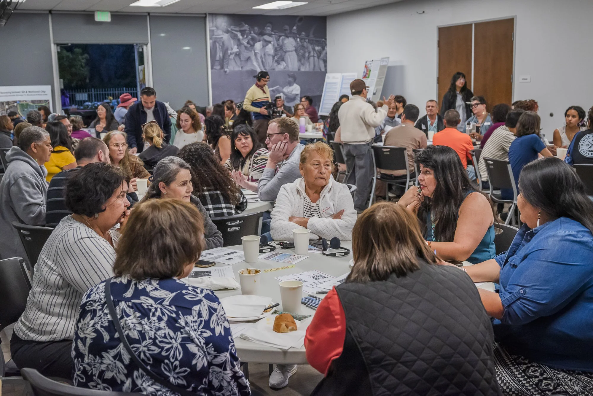 People sitting around tables in a large indoor room, engaging in a meeting or discussion, with some listening, some speaking, and others observing. There are informational posters and a mural or photograph on the wall in the background.
