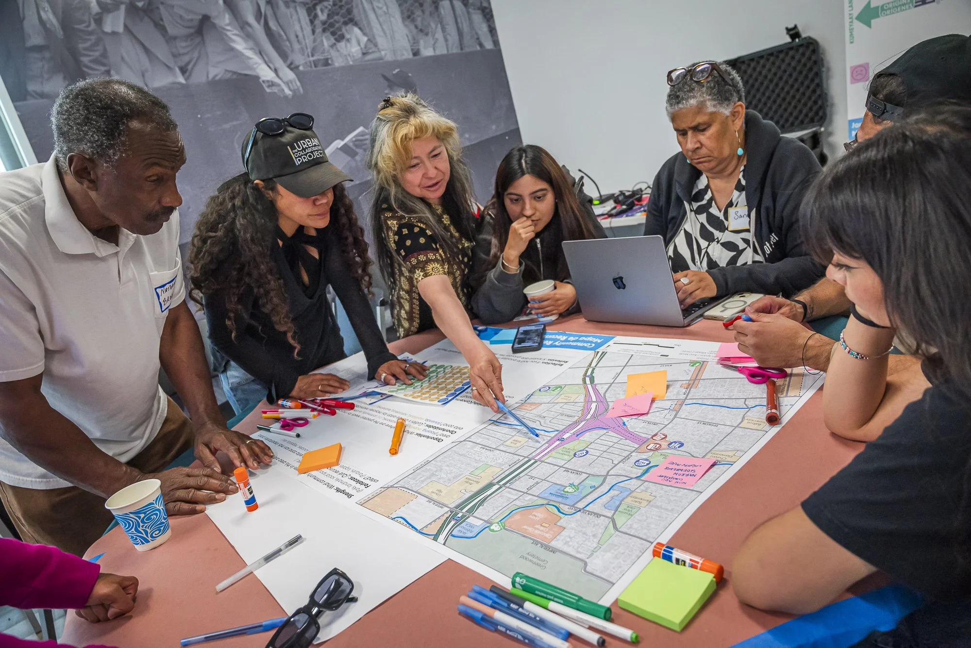 A diverse group of people gathered around a table collaborating on a community planning project, examining a large map with colorful markings, markers, and sticky notes.