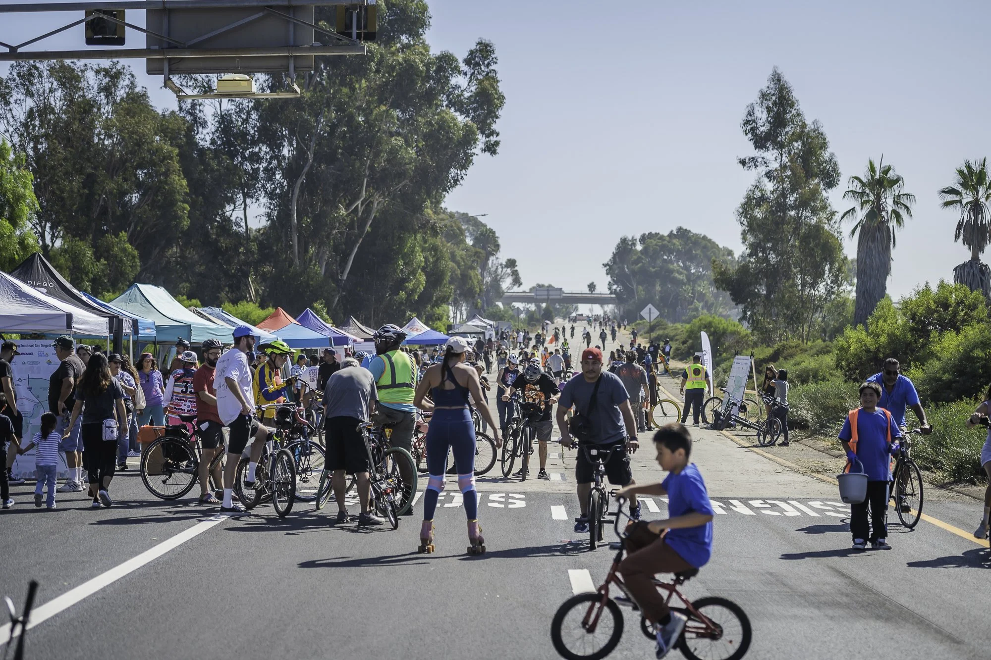 Many people gathered at a biking event on a sunny day, with tents along the side of the road and children riding bikes in the foreground. Trees and a highway overpass are visible in the background.