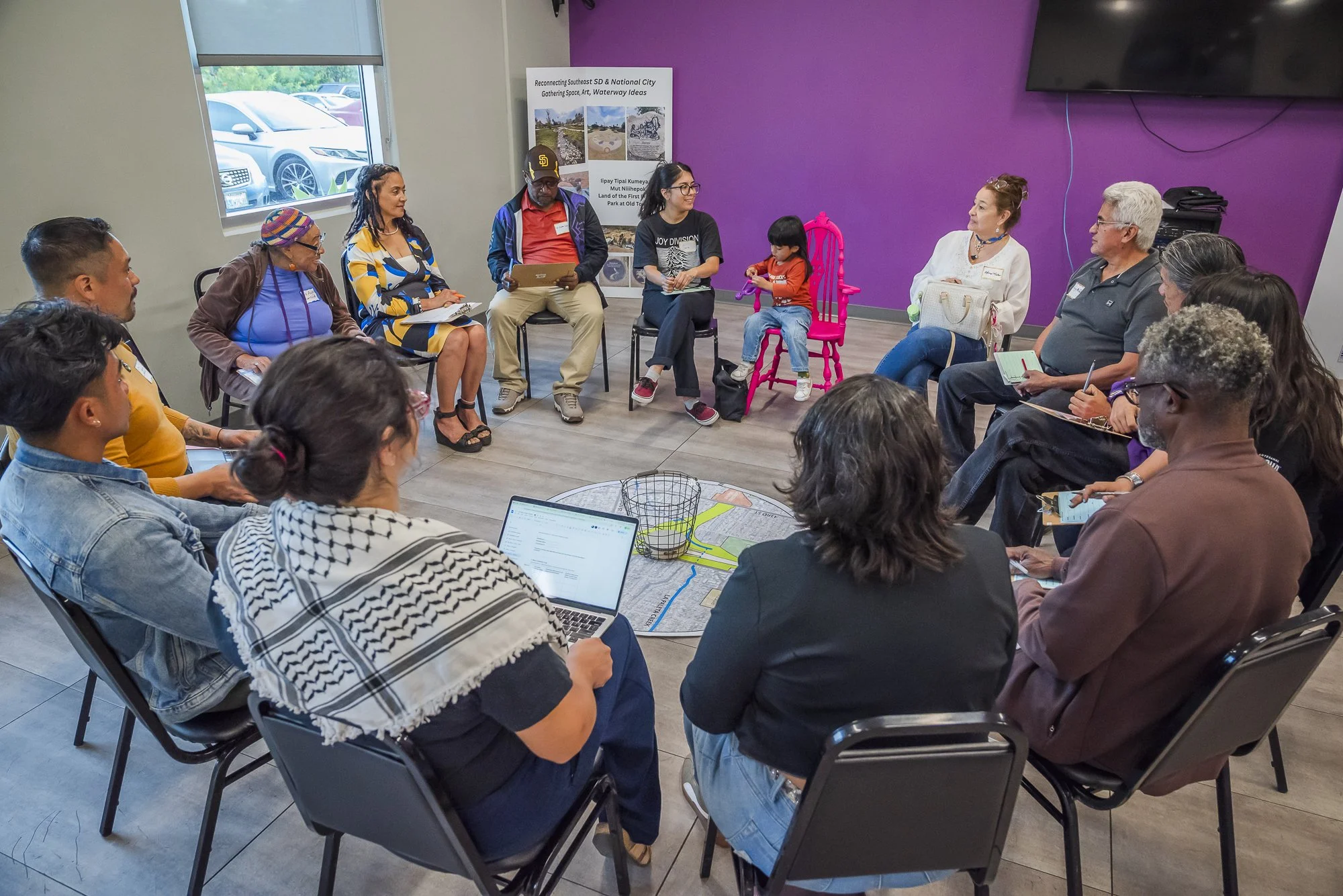 A diverse group of people gathered in a circle for a meeting in a room with purple walls and a large window. Some are holding notebooks, tablets, or pens, and a little girl is sitting in a pink chair.