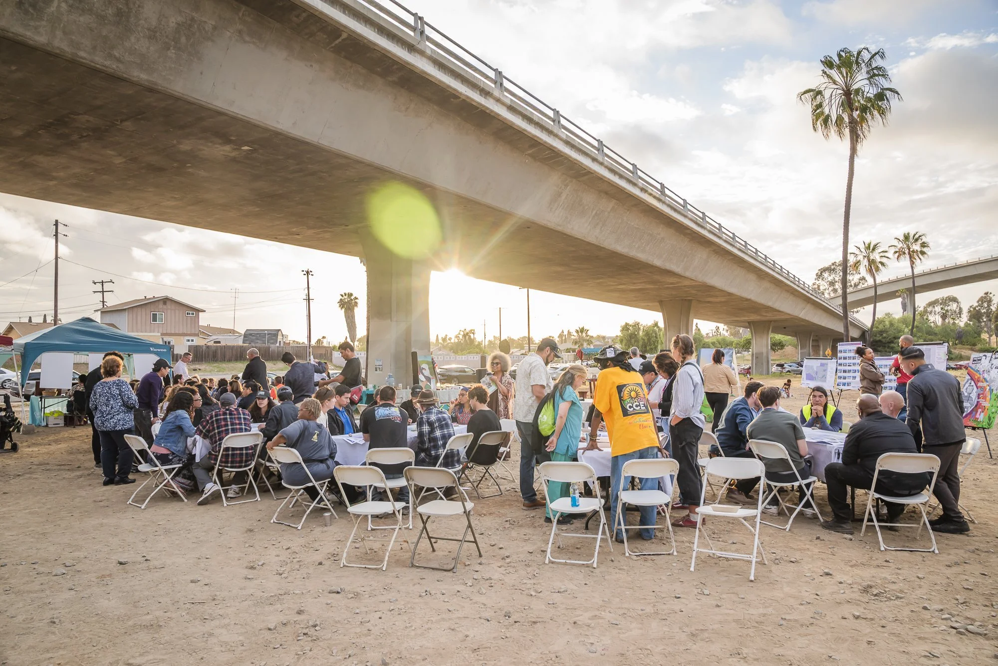 People gathered outdoors under a bridge, participating in an event with tables, chairs, tents, and informational displays. The setting appears to be in a sunny, semi-urban area with palm trees.