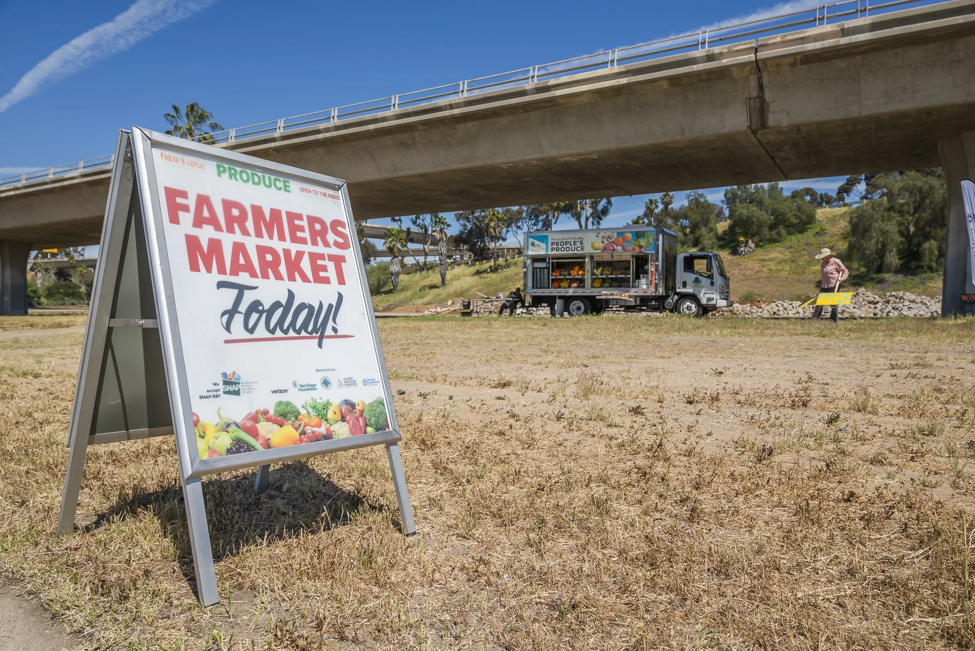 A signboard advertising a farmers market with colorful fruits and vegetables, set up outdoors under a bridge with a food truck and a person in a hat in the background.