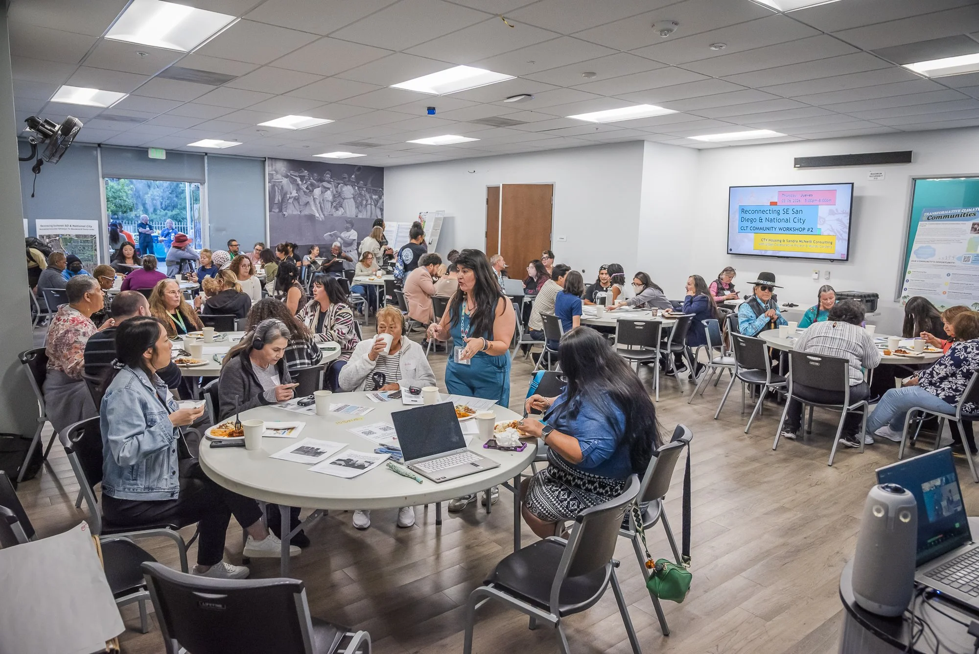 A large room filled with people attending a community workshop or meeting. Several round tables with chairs are occupied by individuals eating, drinking, or using laptops. A woman in a blue outfit is walking among the tables. A wall-mounted display s