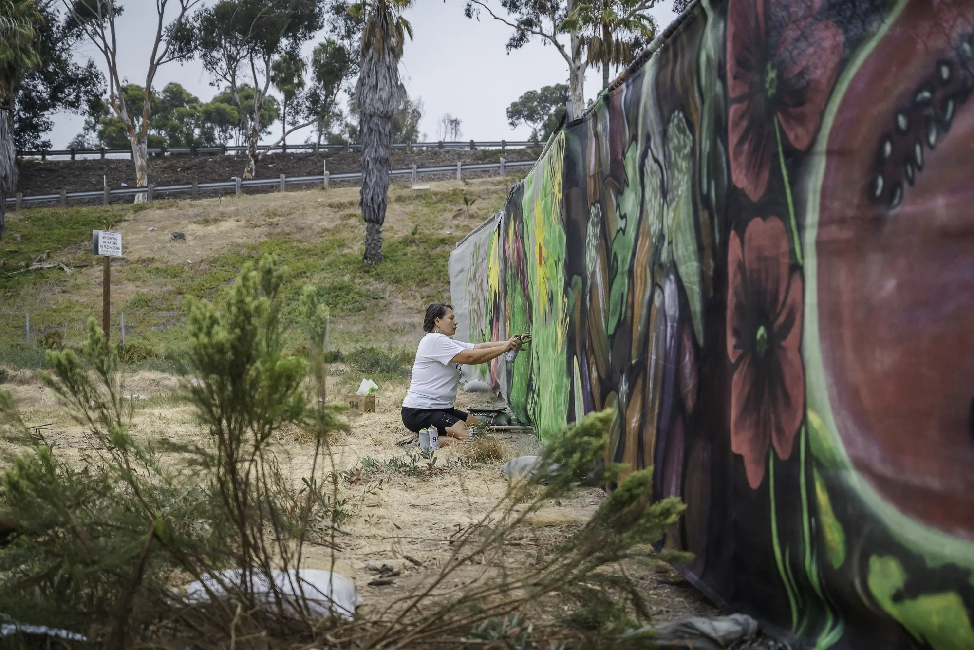 Woman painting a large outdoor mural with colorful floral and nature designs. She is kneeling on the ground, working on the mural, with trees and a fence in the background.