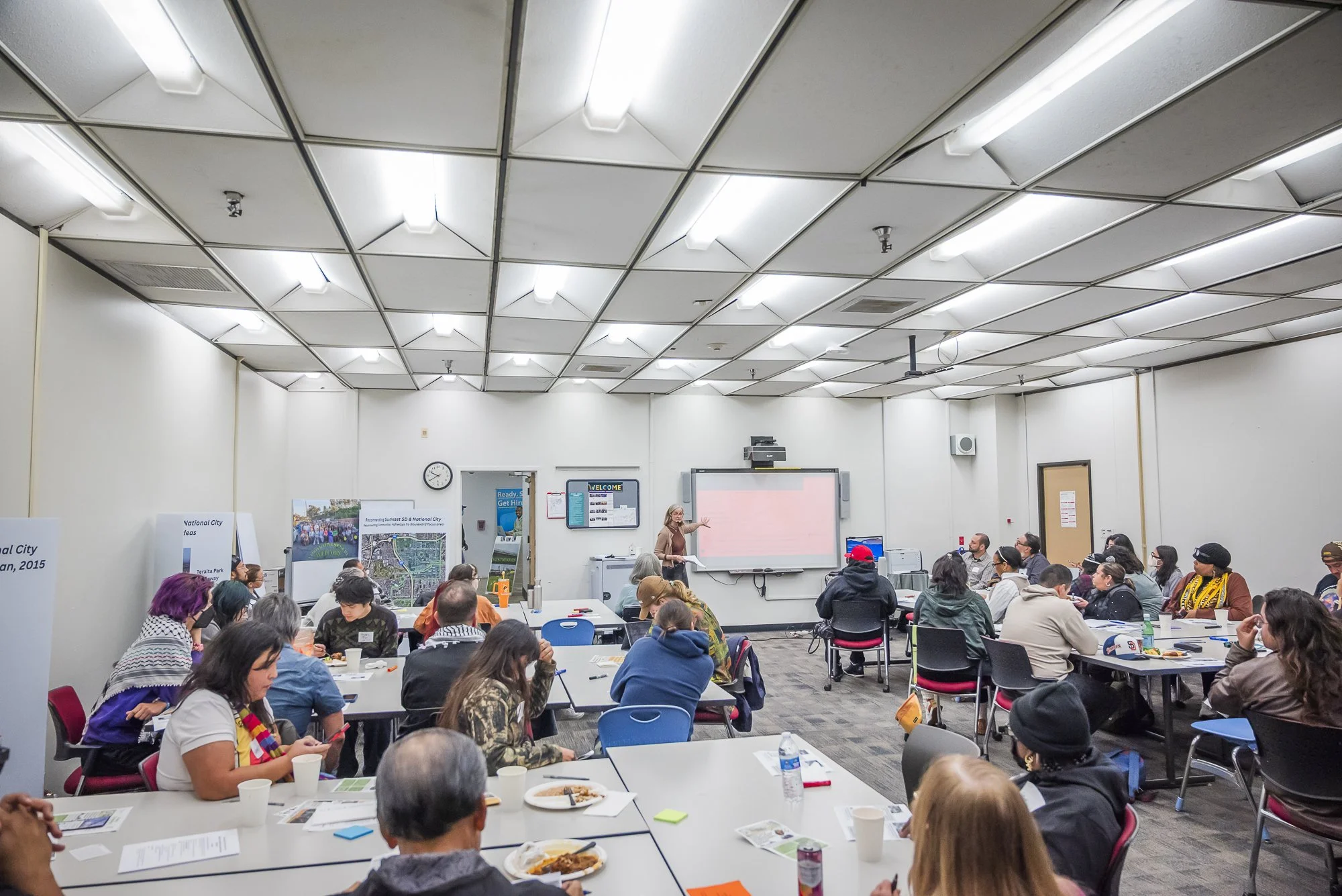 A large group of diverse people seated at tables in a conference room or classroom, listening to a woman presenting at the front of the room. The room has white walls, a whiteboard, and posters, with a ceiling of white panels and fluorescent lighting