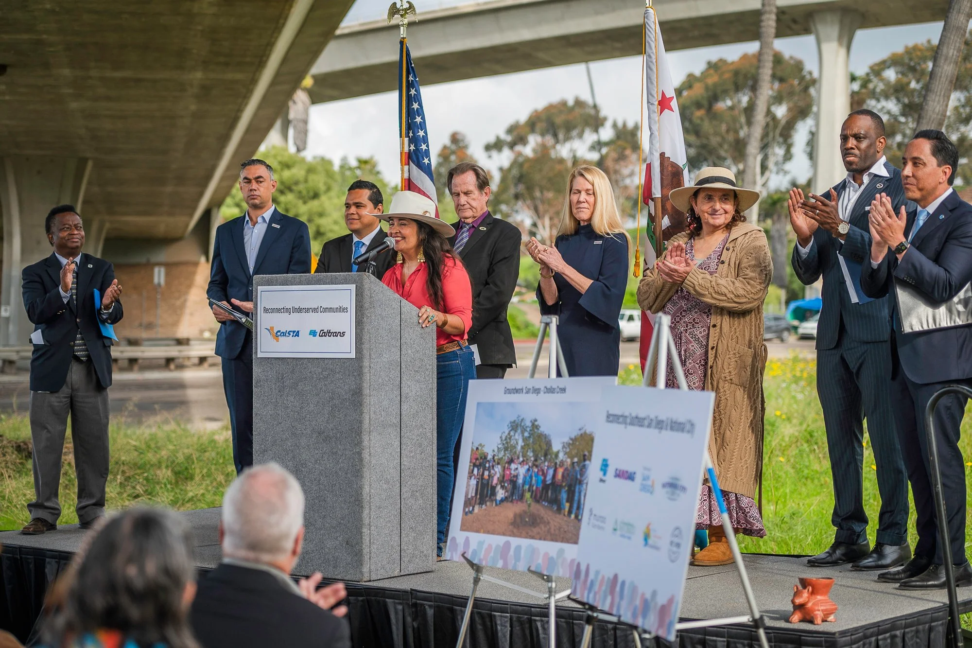 A group of diverse people standing on an outdoor stage, with some clapping and a woman speaking at a podium that reads 'Reconnecting Underserved Communities' under a freeway bridge. Todd Gloria, Toks Omishakin, Tony Tavares, Vicki Estrada
